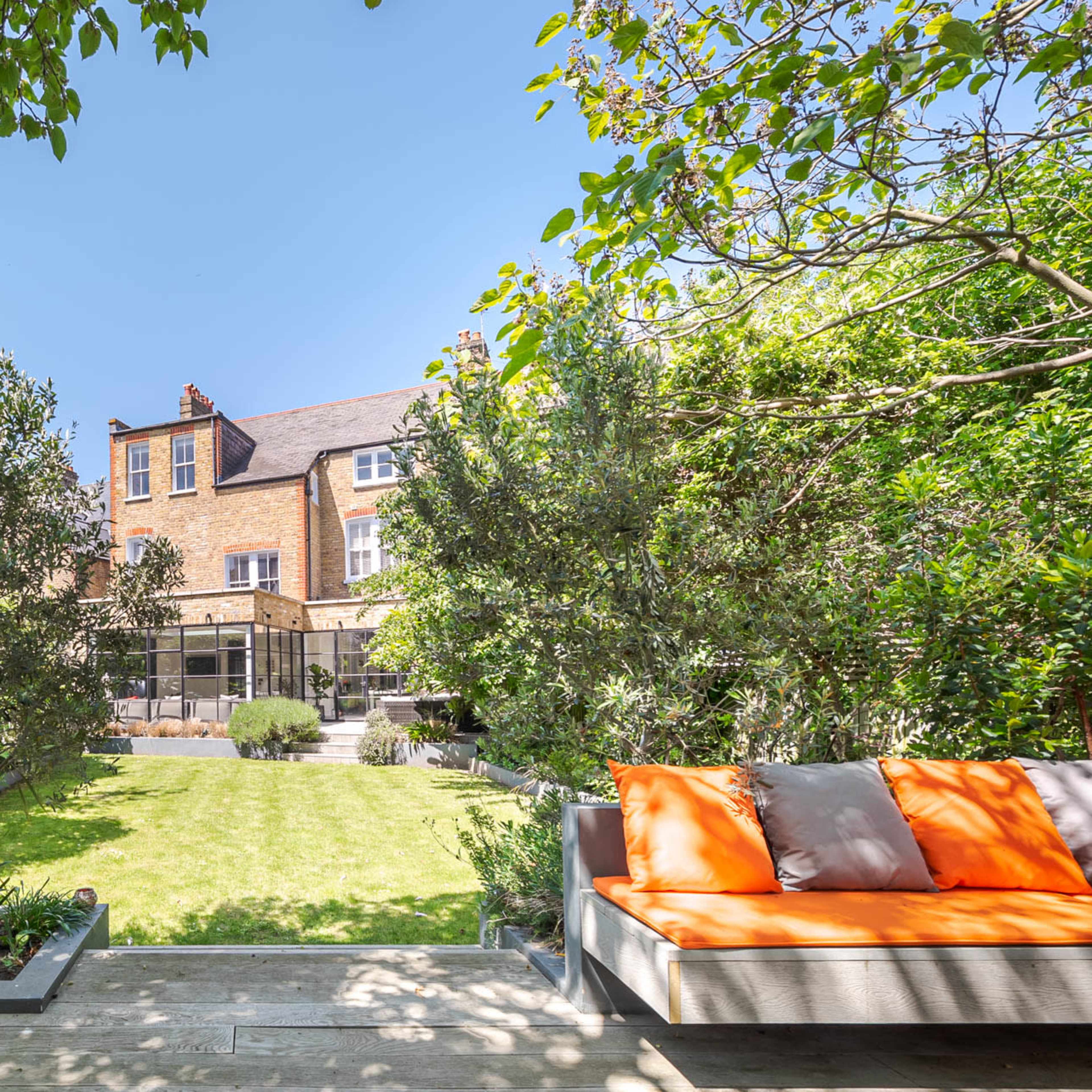 A well-maintained garden features greenery, a manicured lawn, and seating with bright cushions, set against a brick house in the background.