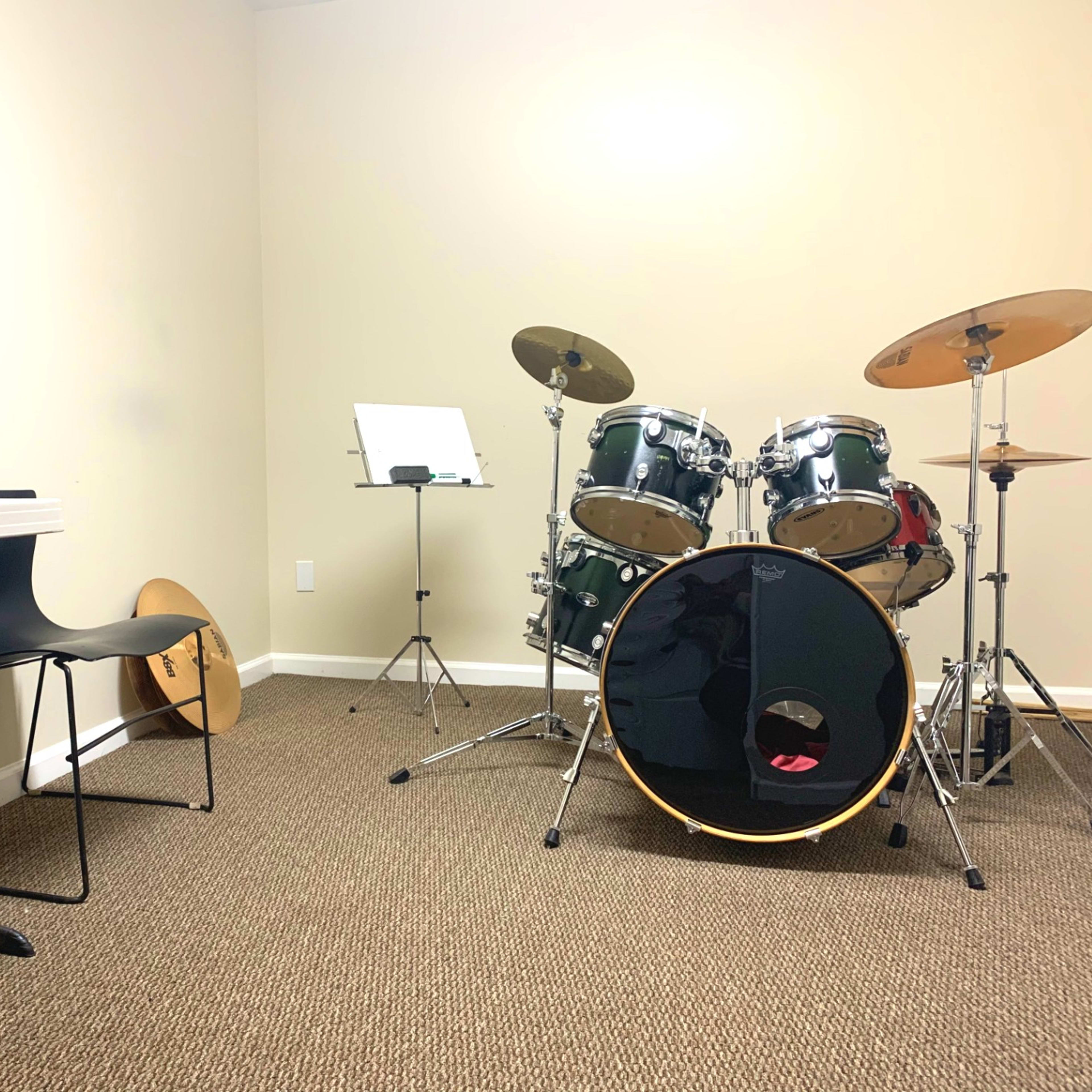 A music practice room features a drum set, a folding chair, and a music stand against beige walls.