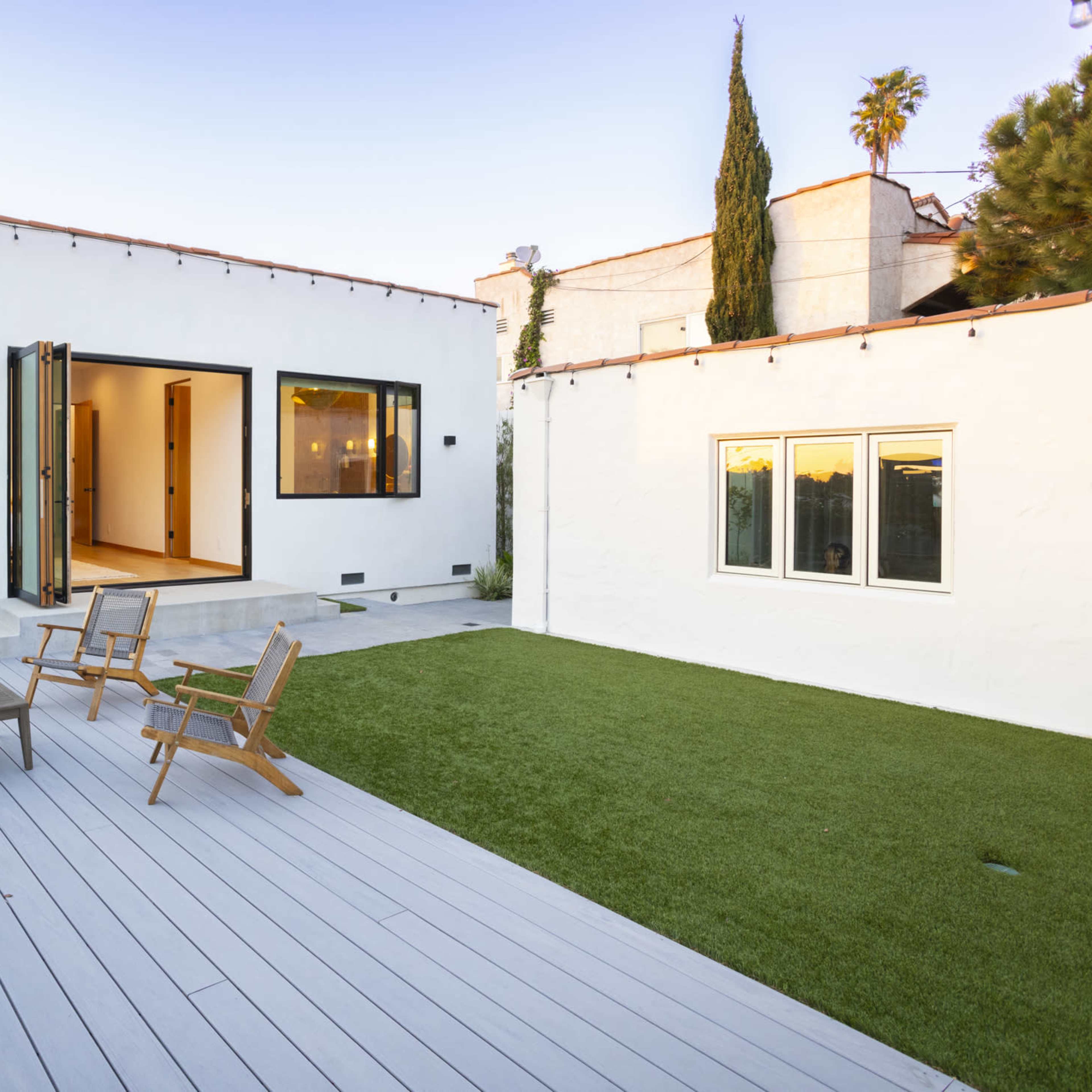 A modern backyard with a wooden deck, two chairs, a small table, and a manicured lawn adjacent to a white house.