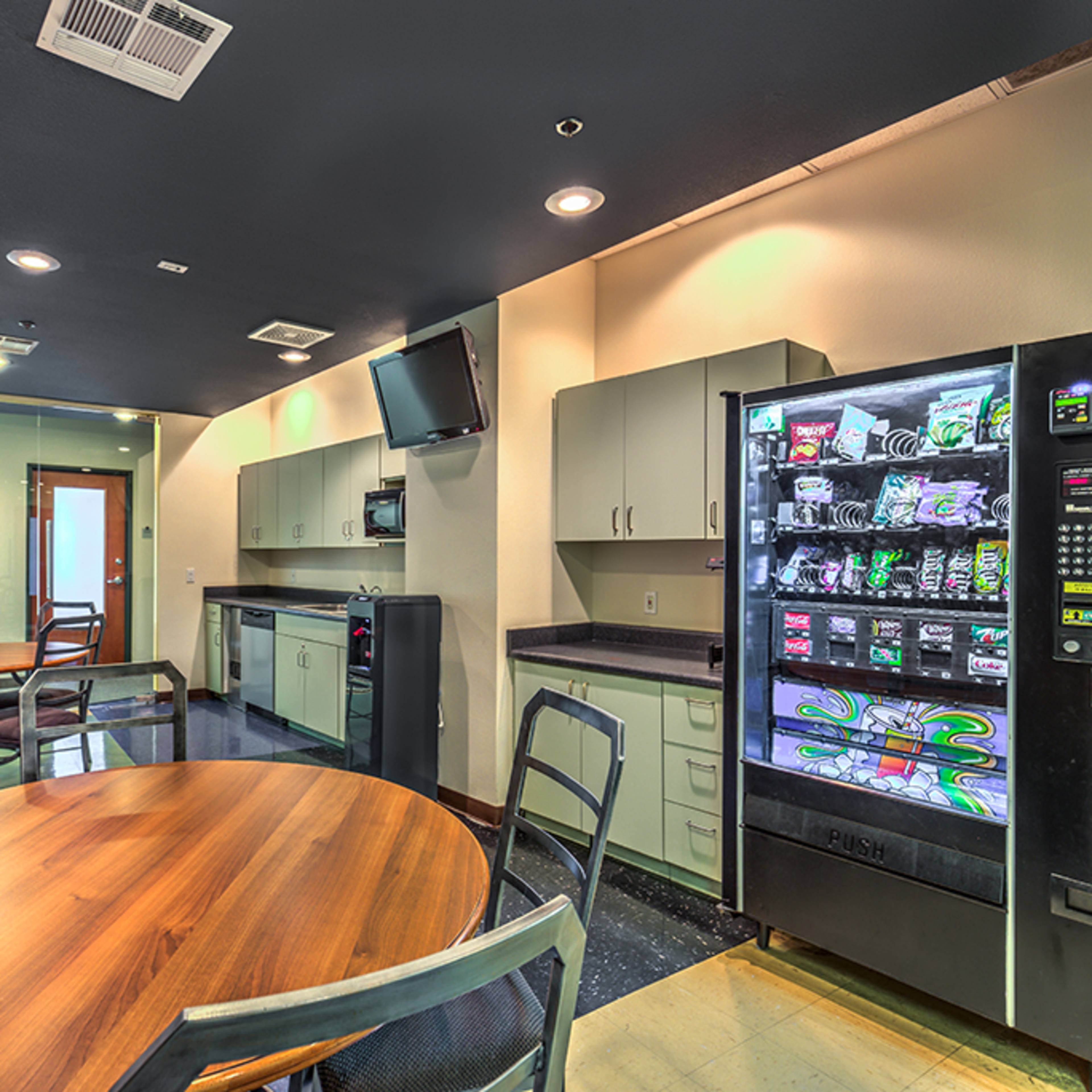 A break room features a wooden table, metal chairs, a vending machine, a refrigerator, and a flat-screen TV on the wall.