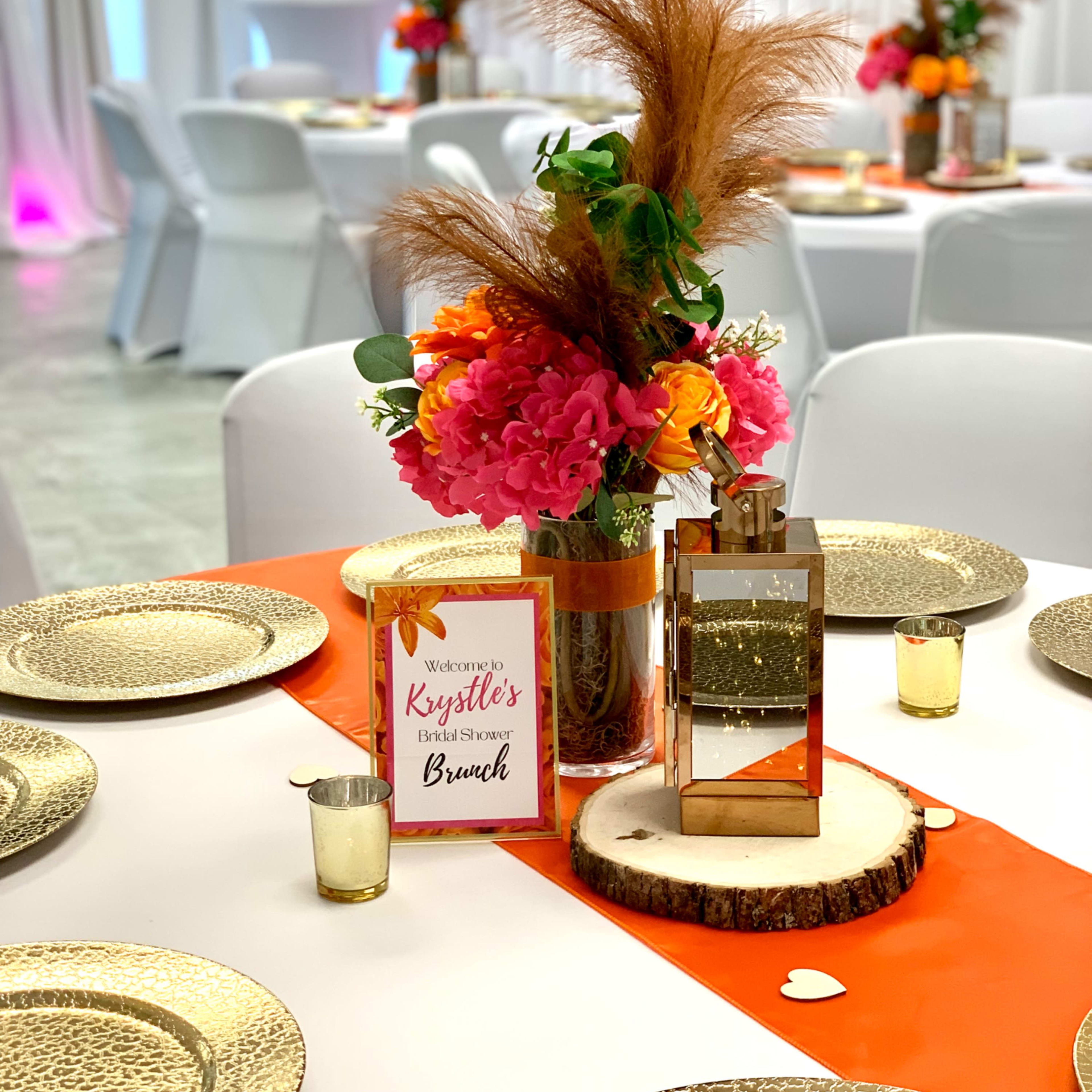 A decorated dining table features a floral centerpiece with pink and orange flowers, a mirrored lantern, and a welcome sign for a brunch event.
