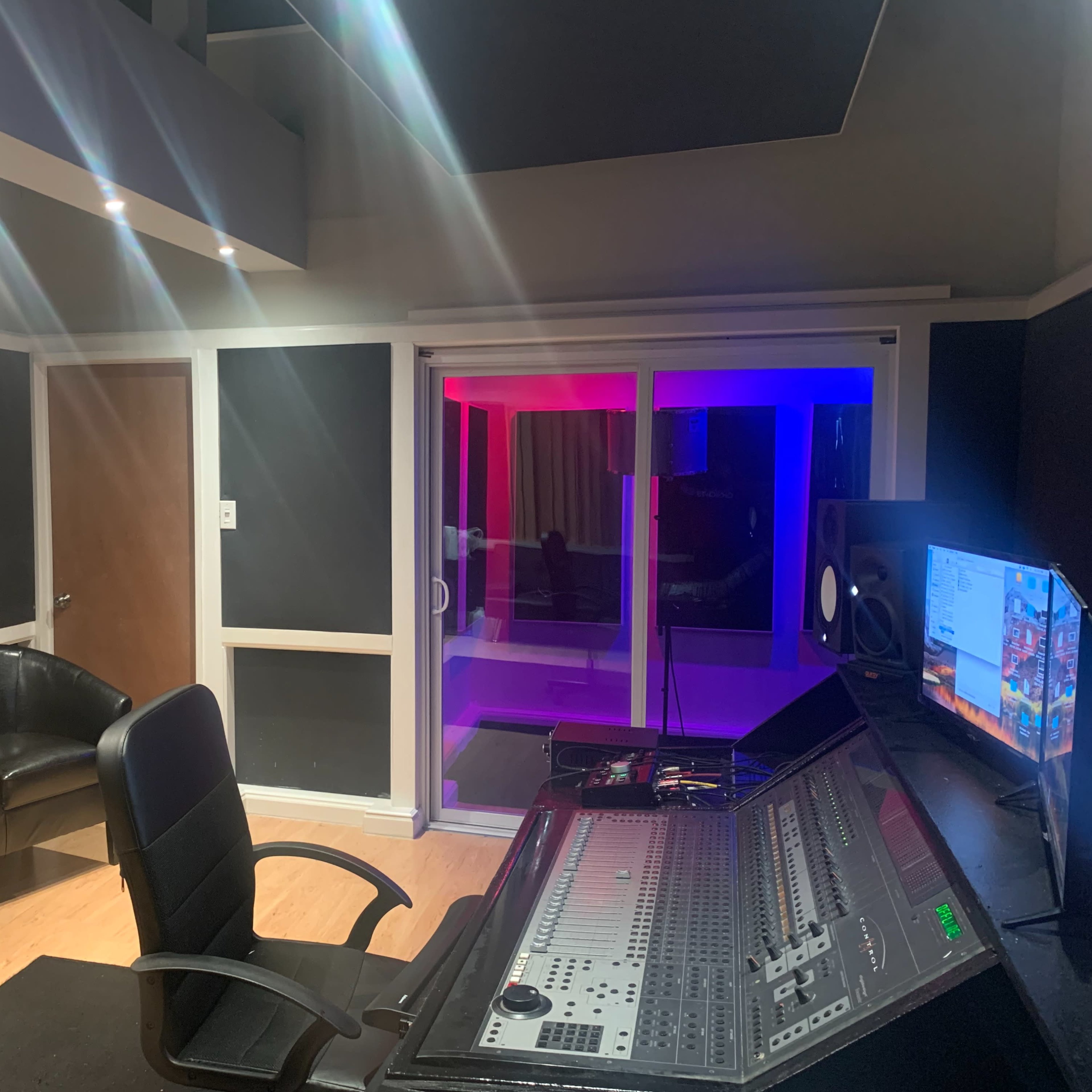 A music studio with a mixing console, a black office chair, a gray couch, and a glass-paneled room illuminated with colored lights.