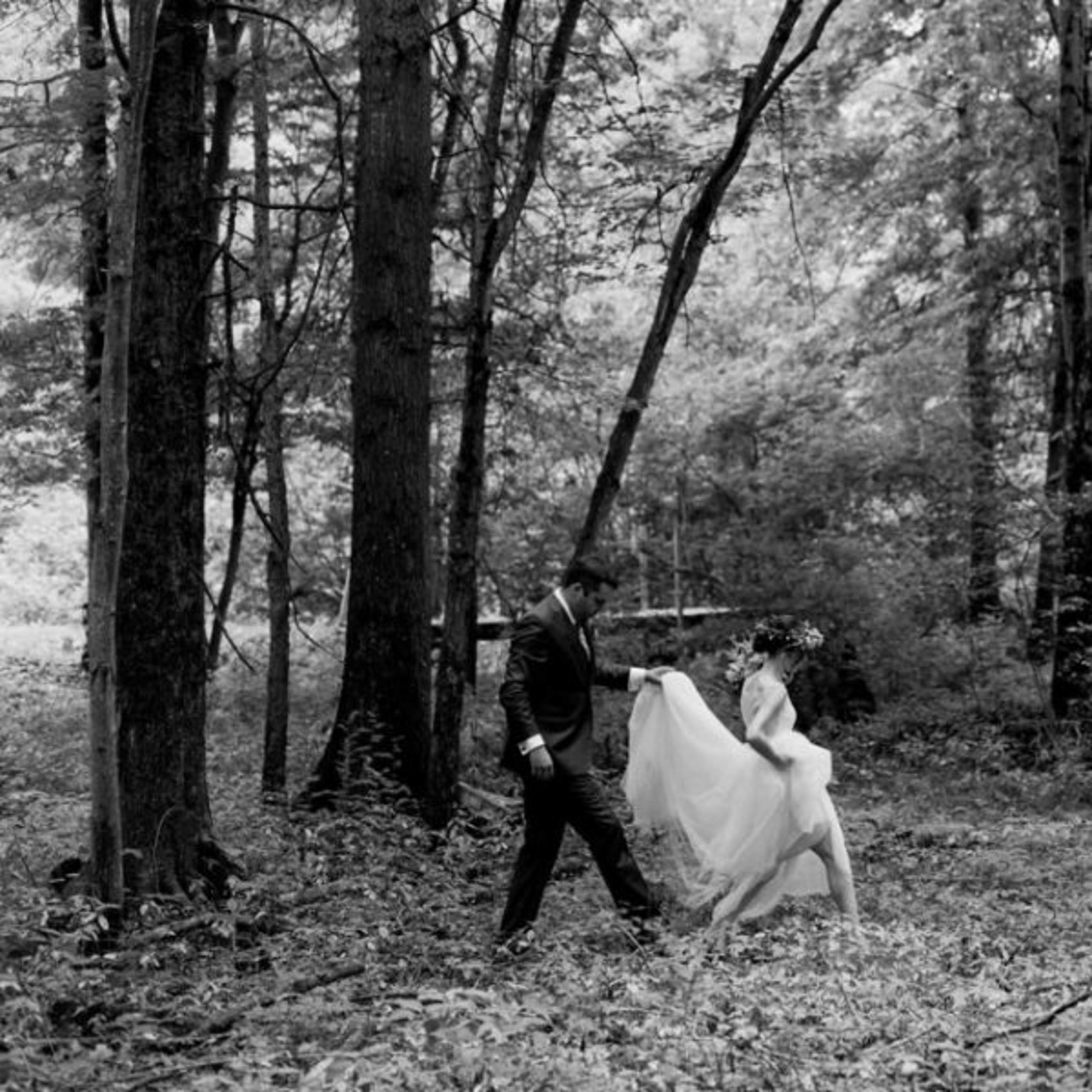 A man in a suit walks with a woman in a wedding dress through a forested area.