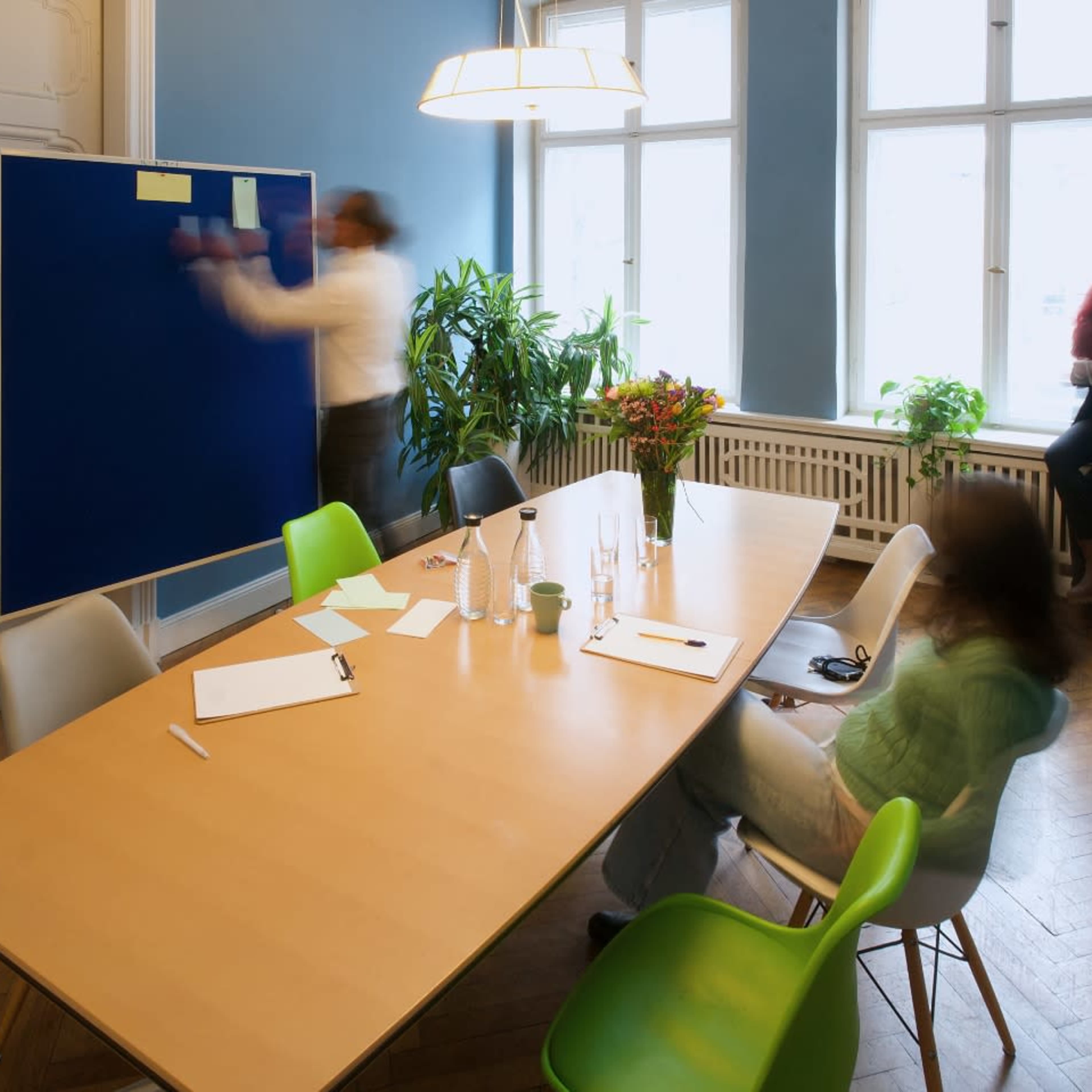 A group of people is engaged in a meeting around a wooden table in a brightly lit room, with one person writing on a blue board while another is seated and observing.