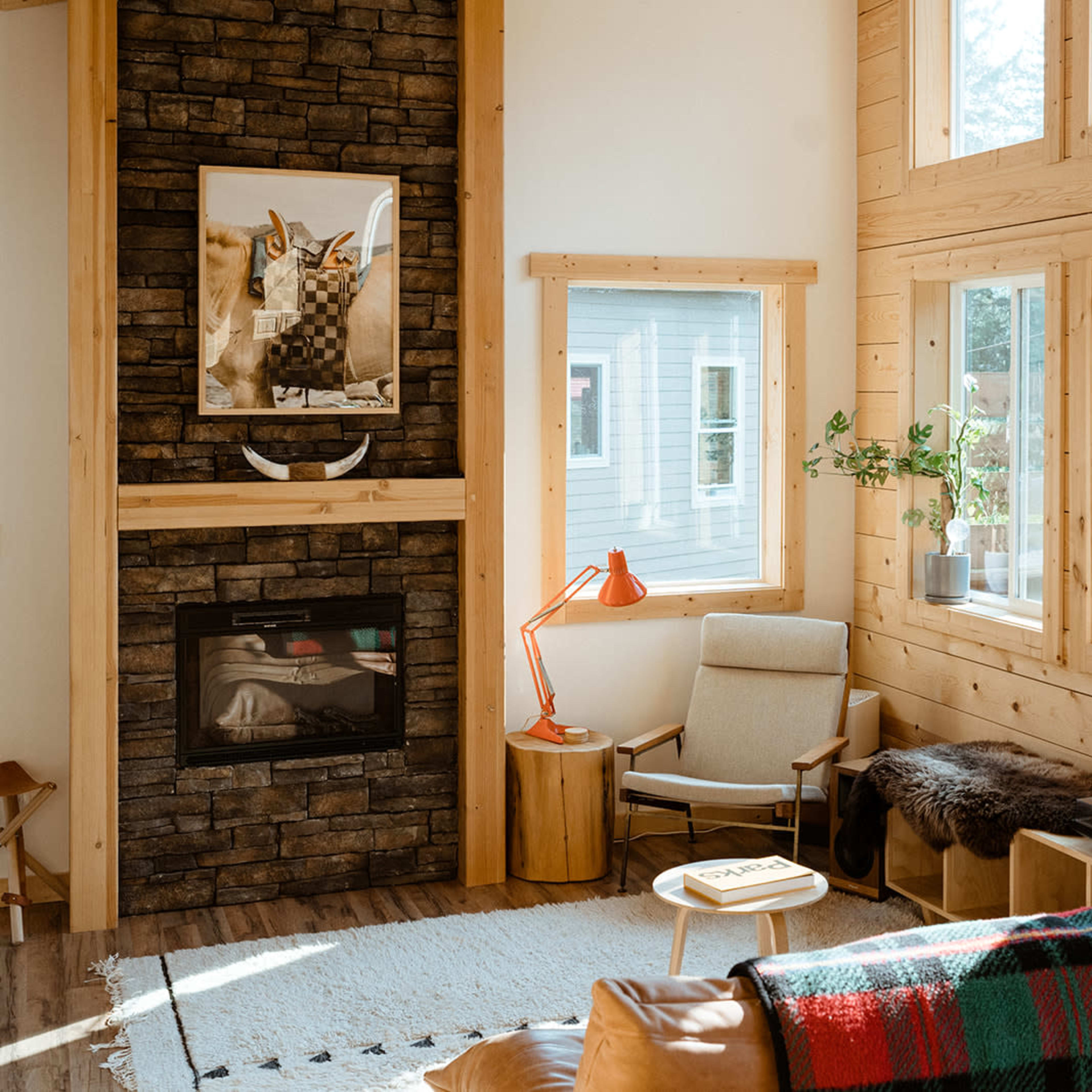 A cozy living room with wood paneling, a stone fireplace, large windows allowing natural light, and minimalistic furnishings including a chair, a side table, and a record player.