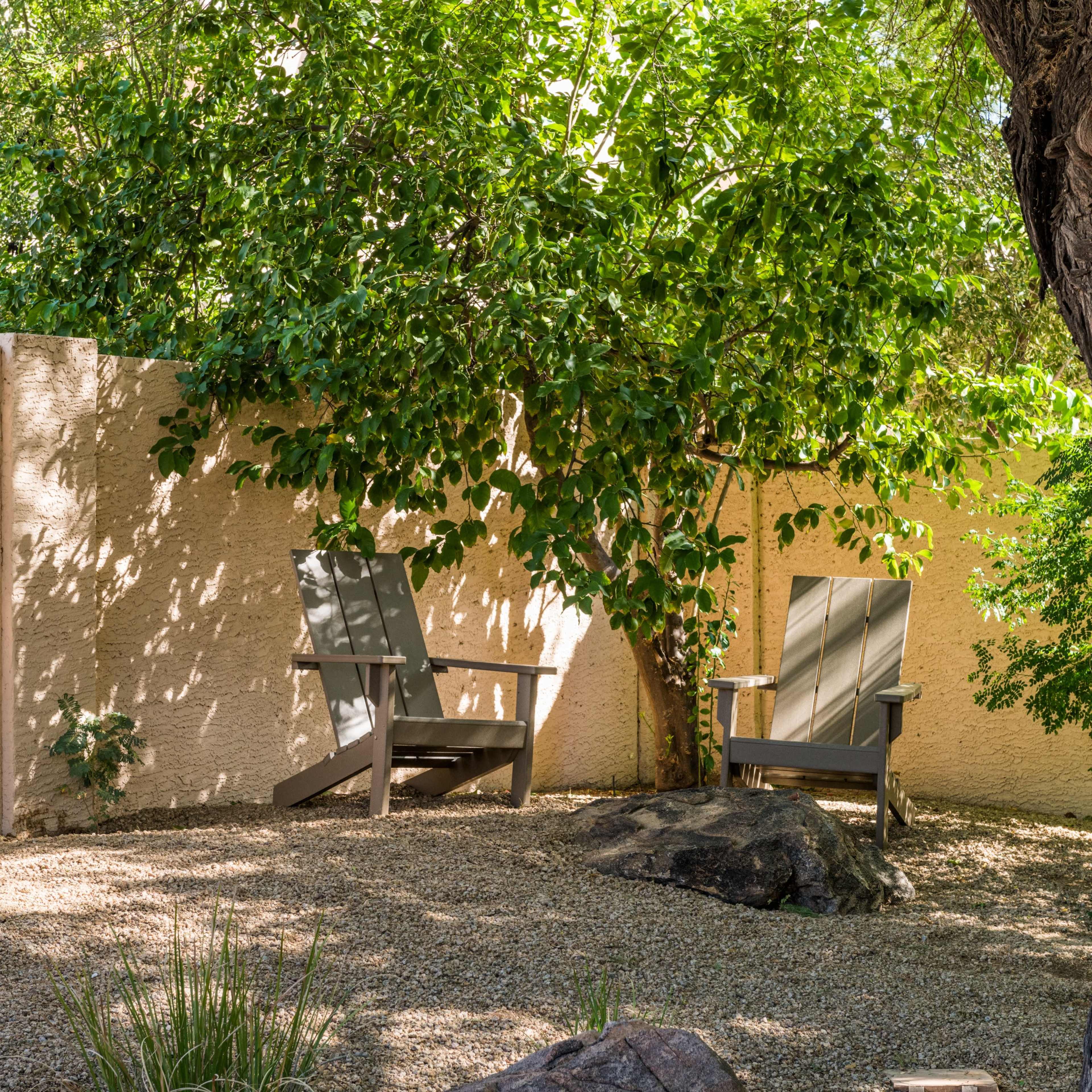 A shaded outdoor area features two wooden chairs beside a stone in a gravel landscape enclosed by a wall and greenery.