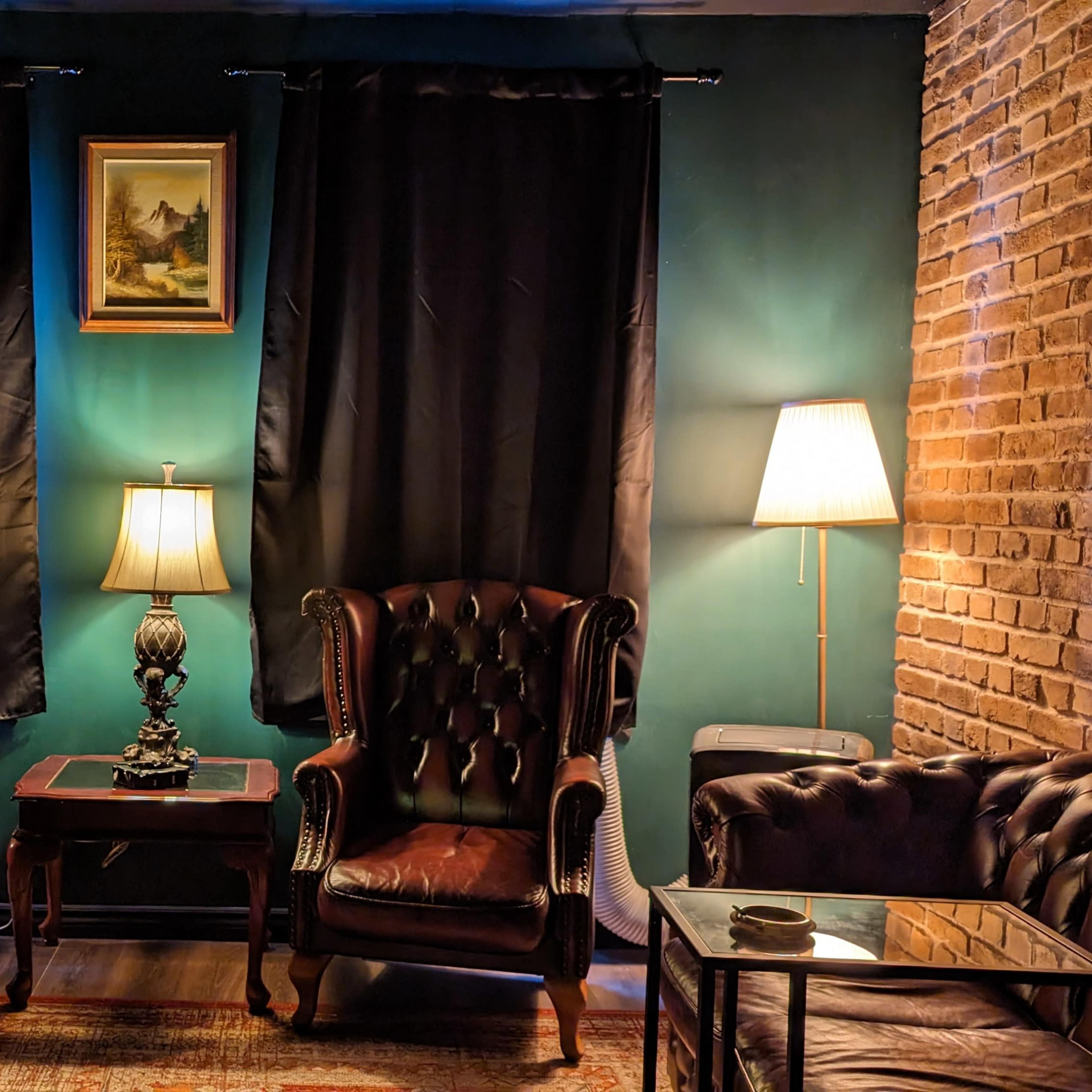 A cozy sitting area features two leather armchairs, a small wooden table, and two floor lamps beside dark curtains and a brick wall.