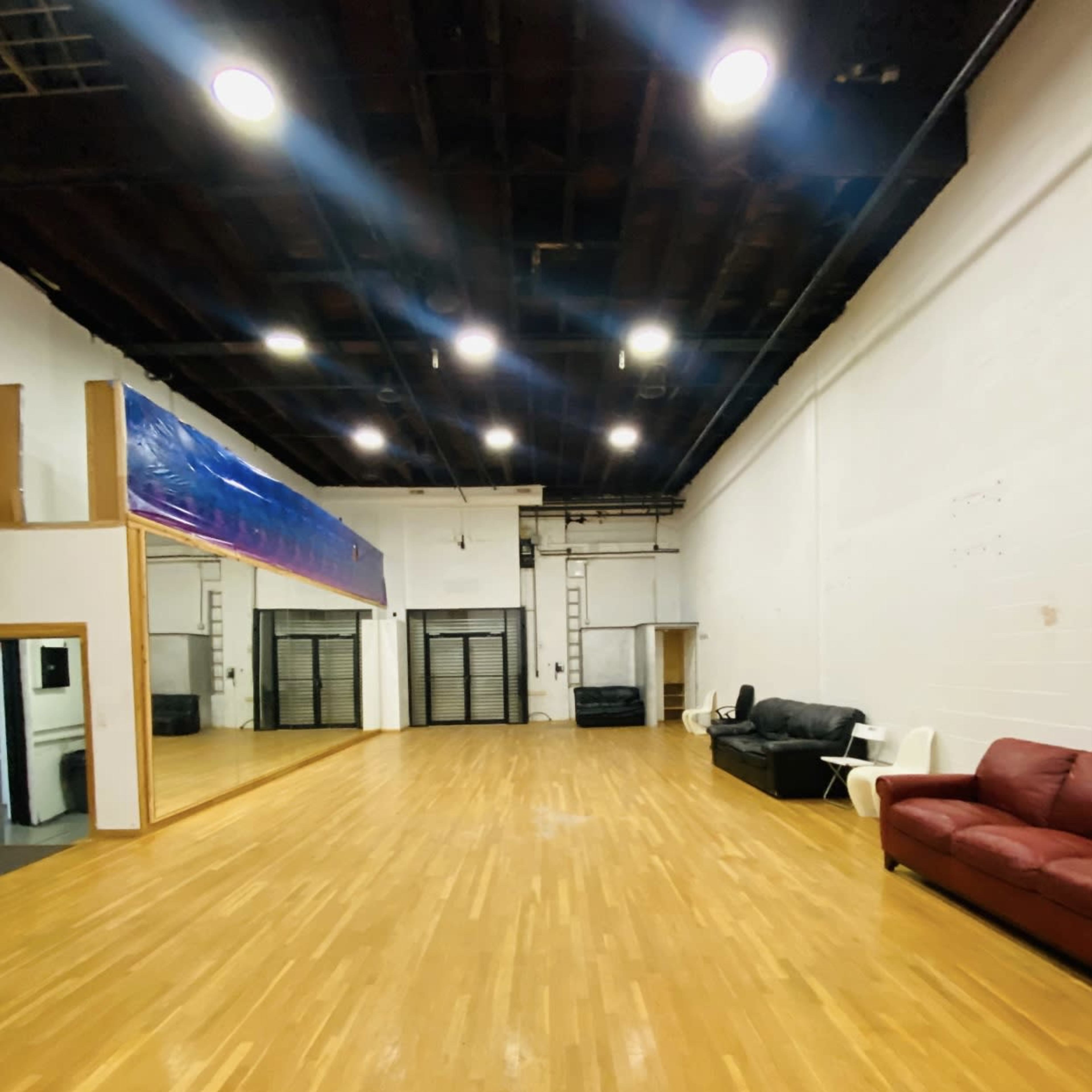 A spacious dance studio with hardwood flooring, large mirrors on one wall, and seating areas comprising two couches.
