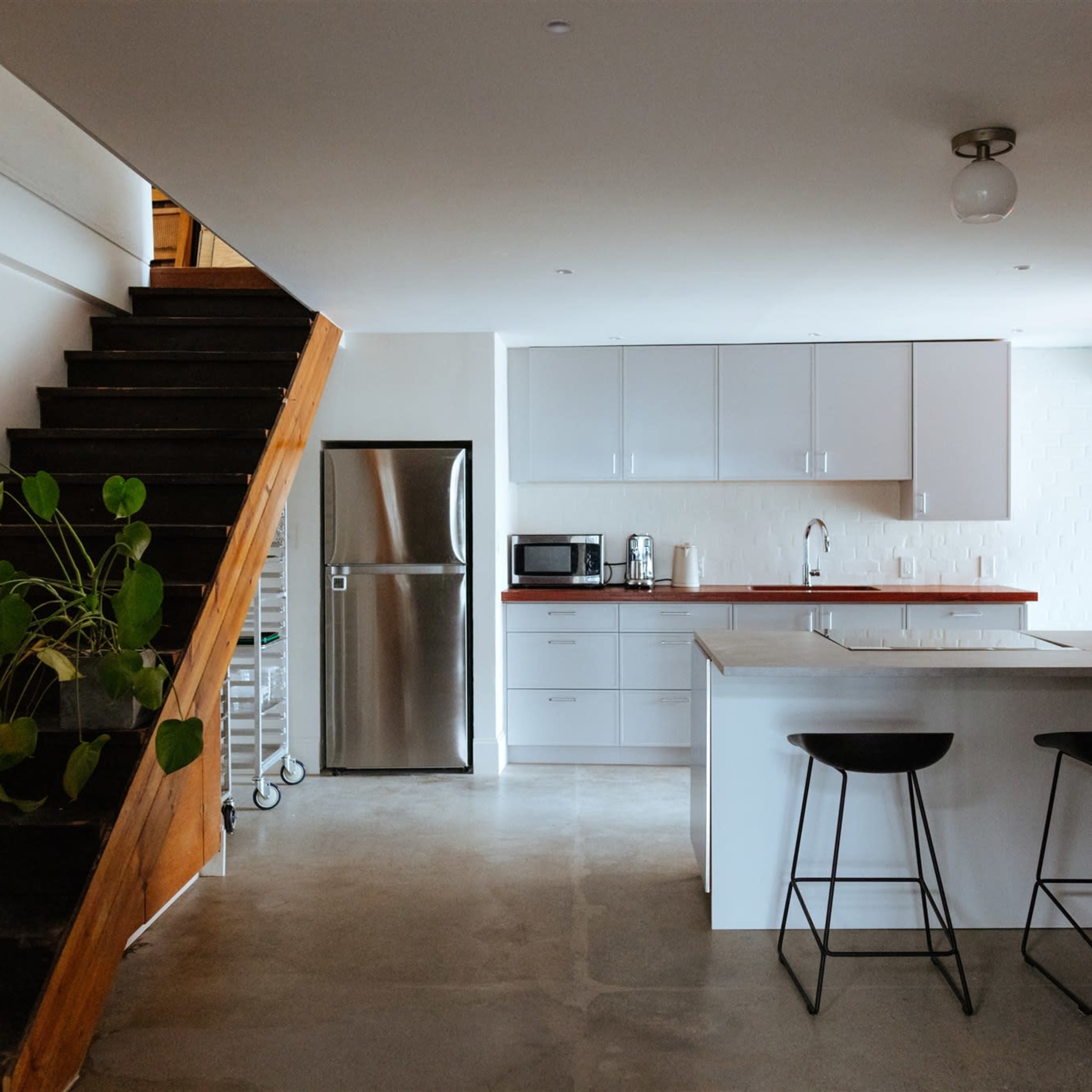 A modern kitchen features a staircase, stainless steel fridge, white cabinets, and black bar stools.
