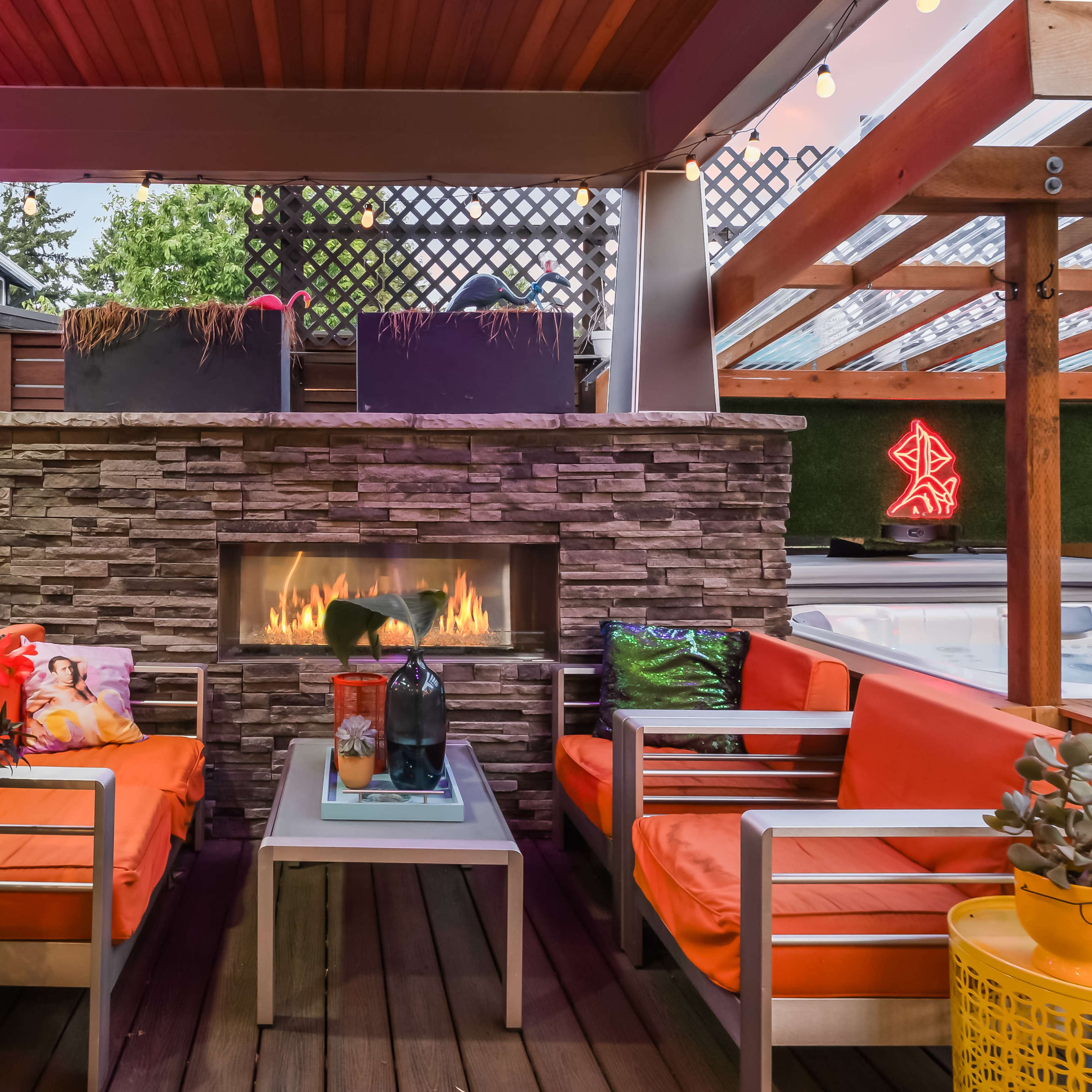 A cozy outdoor seating area with orange chairs, a stone fireplace, and decorative plants under a wooden roof with string lights.