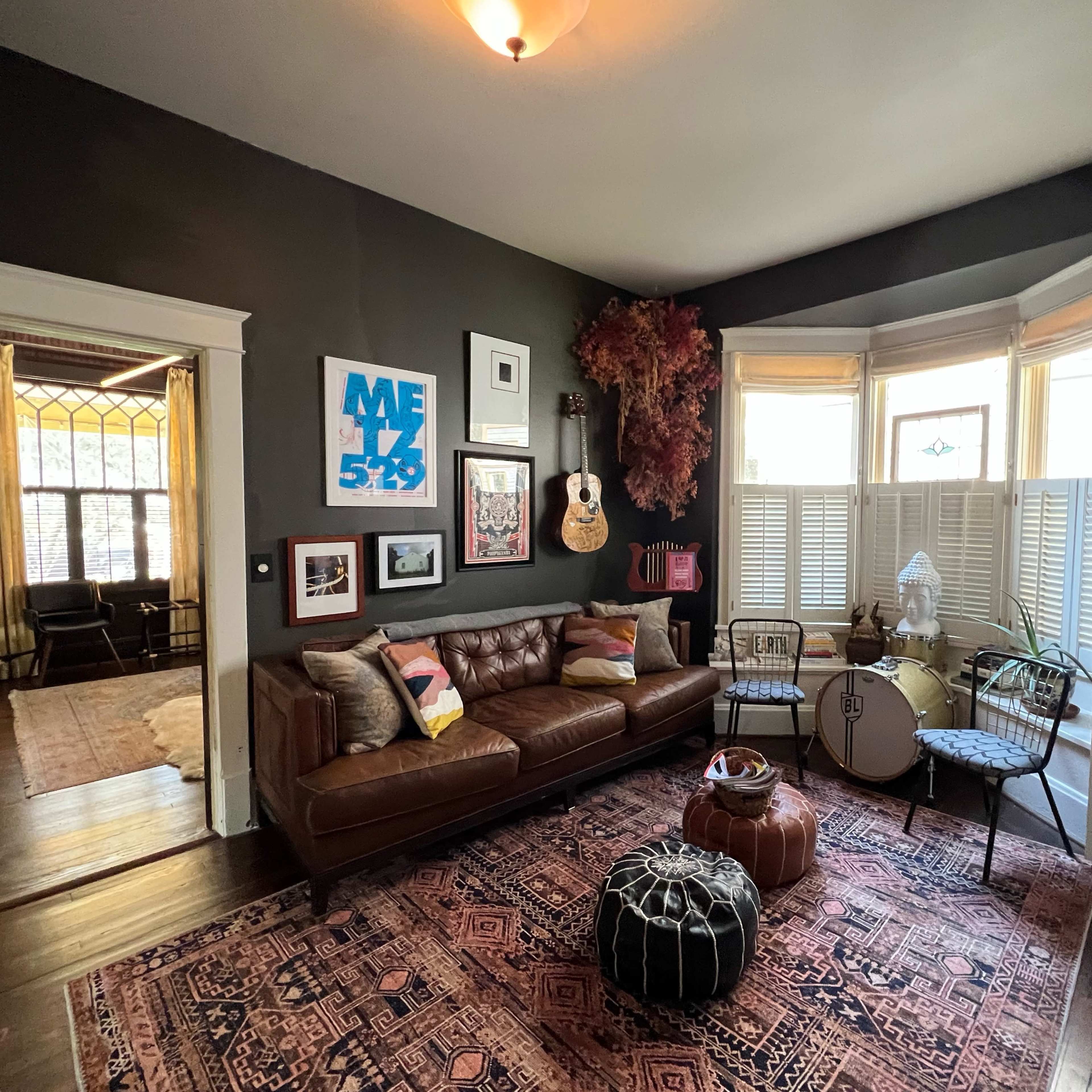 A cozy living room with a brown leather sofa, various framed artworks on the walls, and a large area rug under a round ottoman and decorative chairs near a bay window.