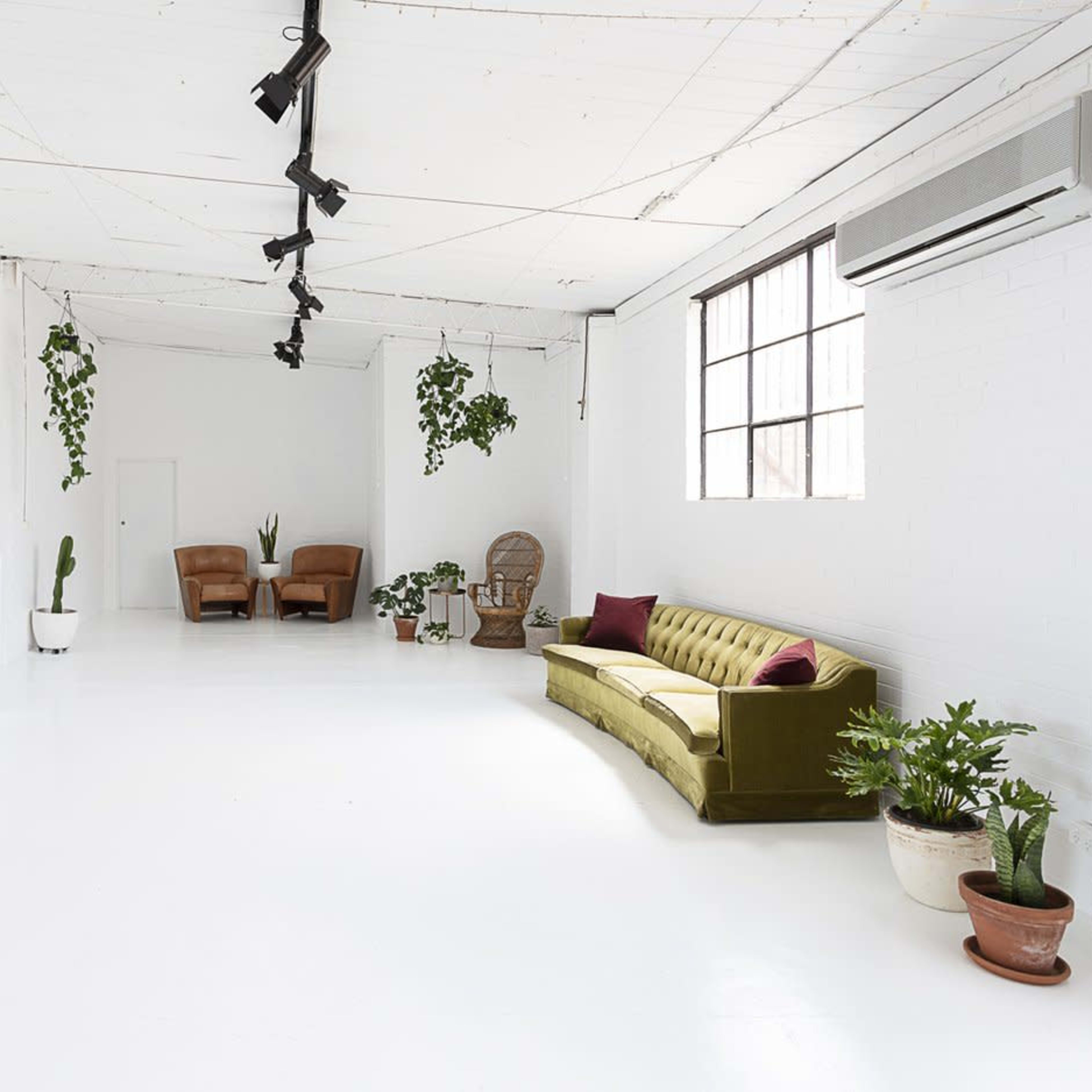 A bright, minimalist studio space featuring a green velvet sofa, two brown chairs, and various plants in pots, with a large mirror and a window.