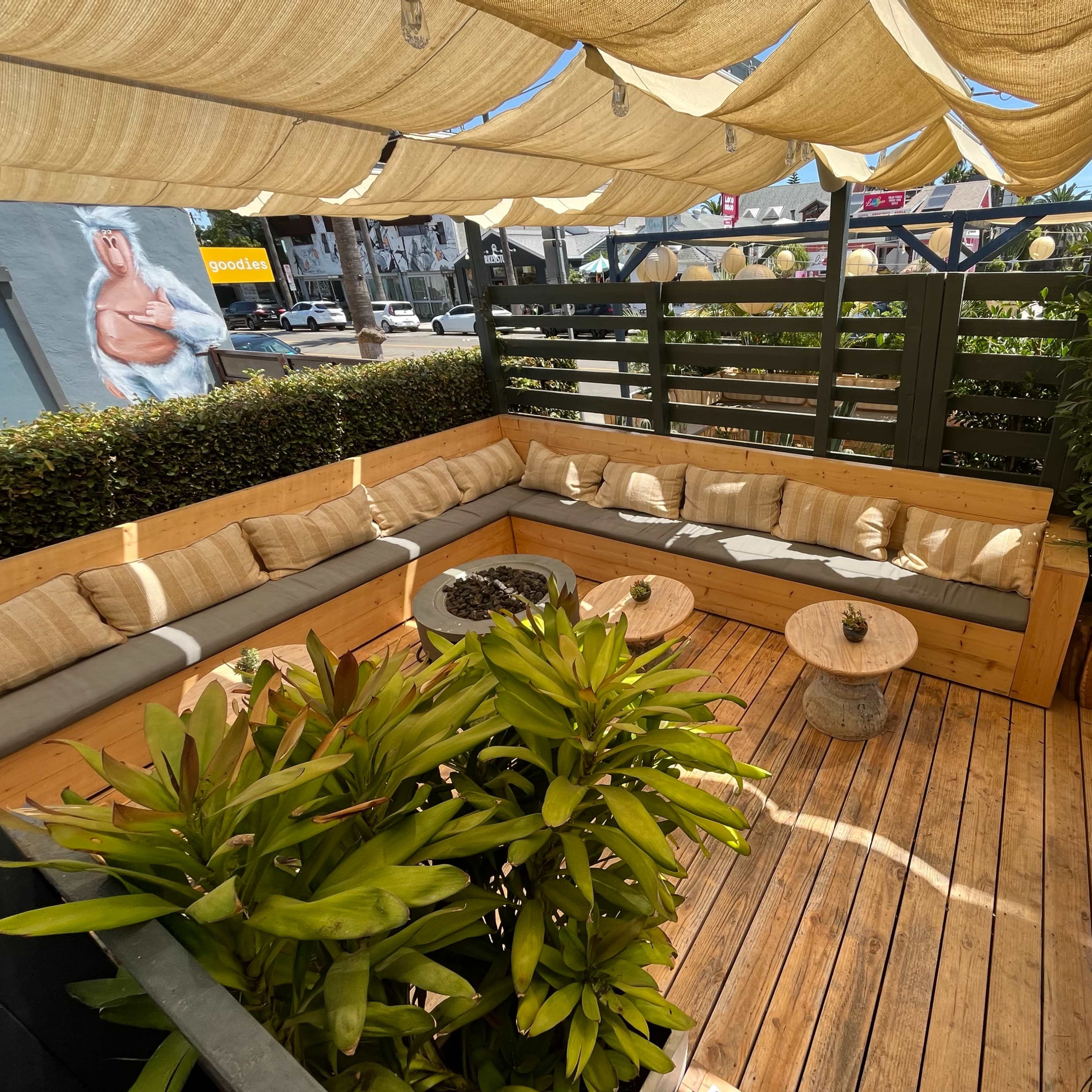 A wooden outdoor seating area is enclosed by low greenery and features a shaded couch with cushions, two small round tables, and potted plants.