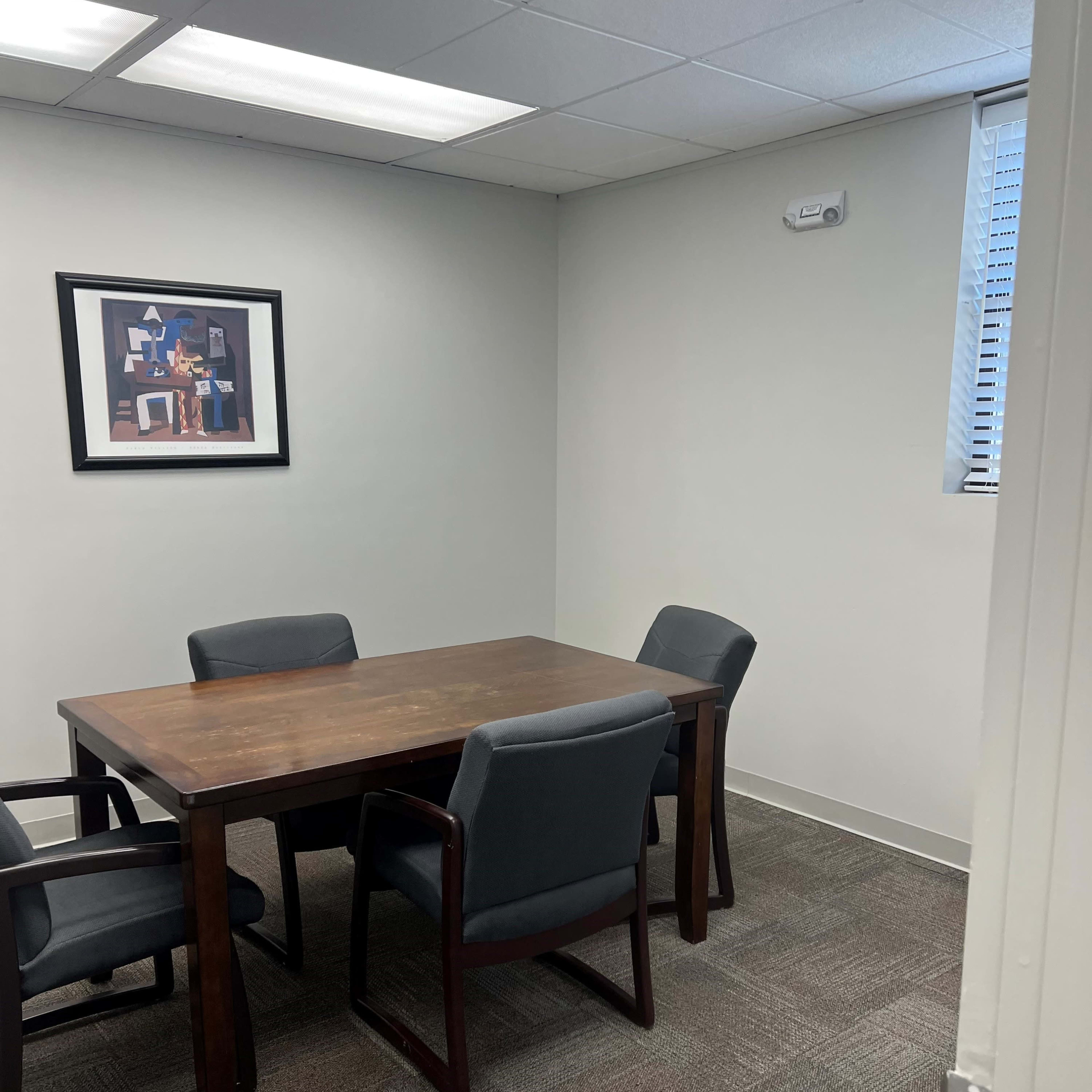 A wooden table with four chairs is placed in a small, well-lit conference room featuring a framed art piece on the wall.