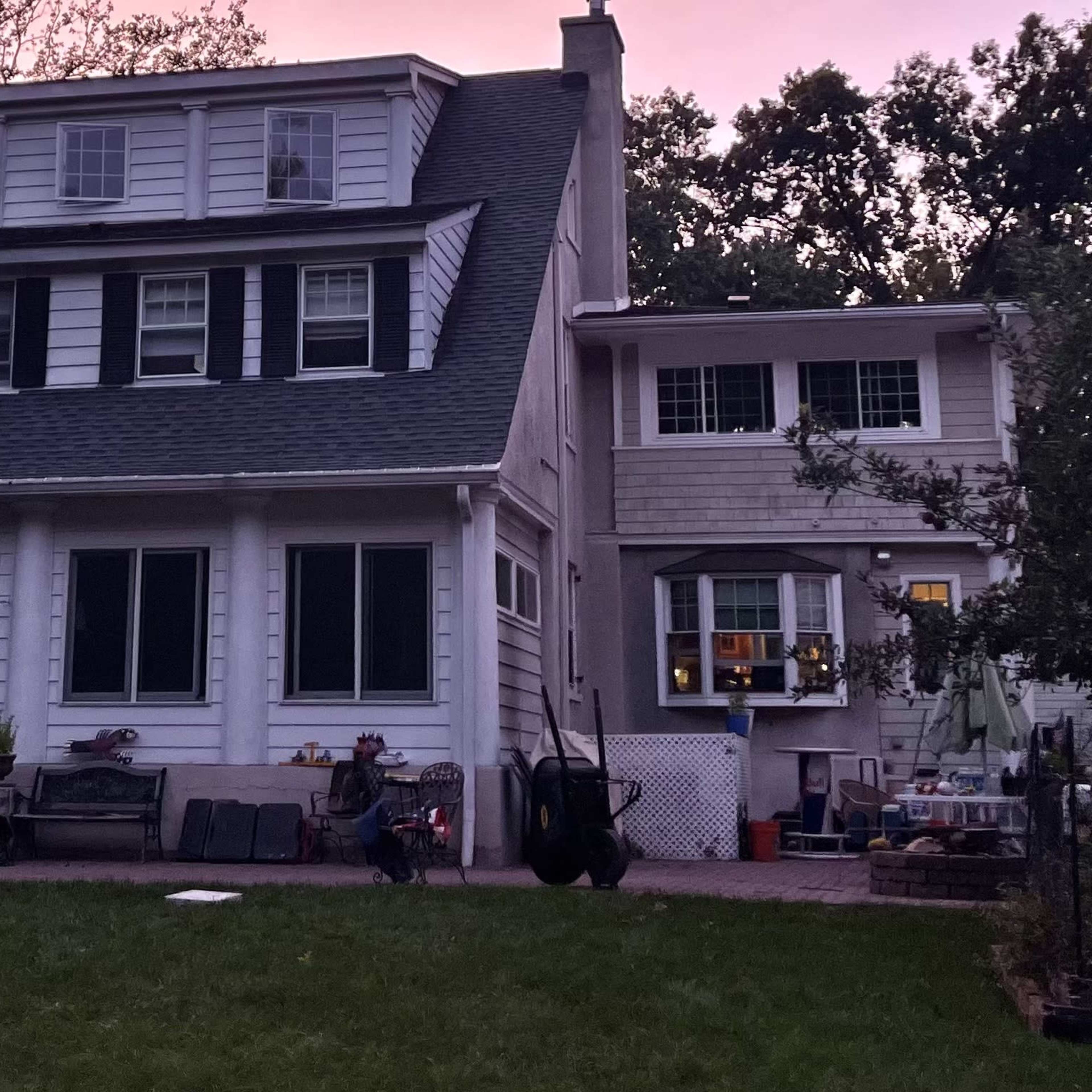A two-story house with multiple windows and a porch is set against a twilight sky, surrounded by trees and a garden area.