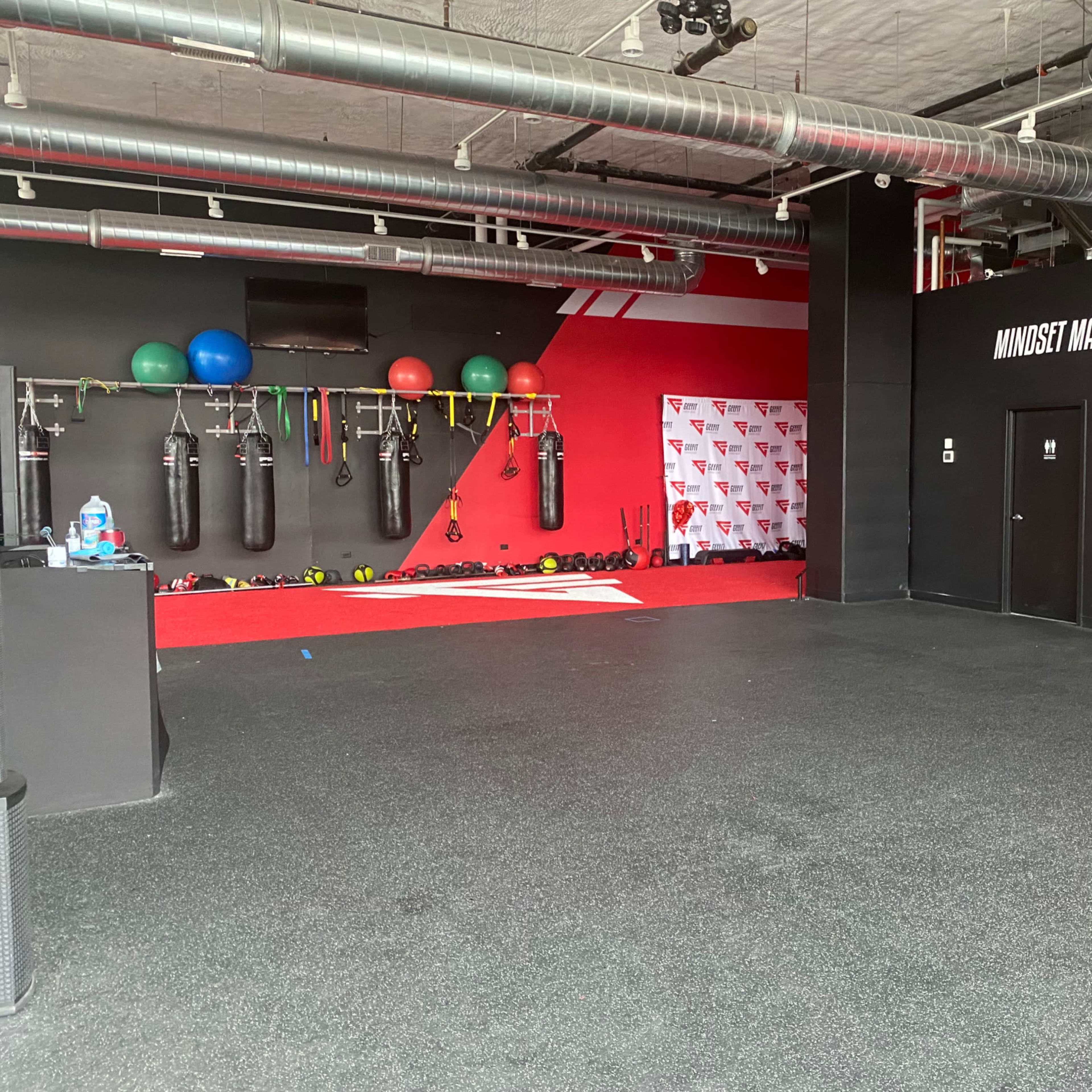 An empty gym with boxing bags, exercise balls, and weight equipment against a red and black backdrop.