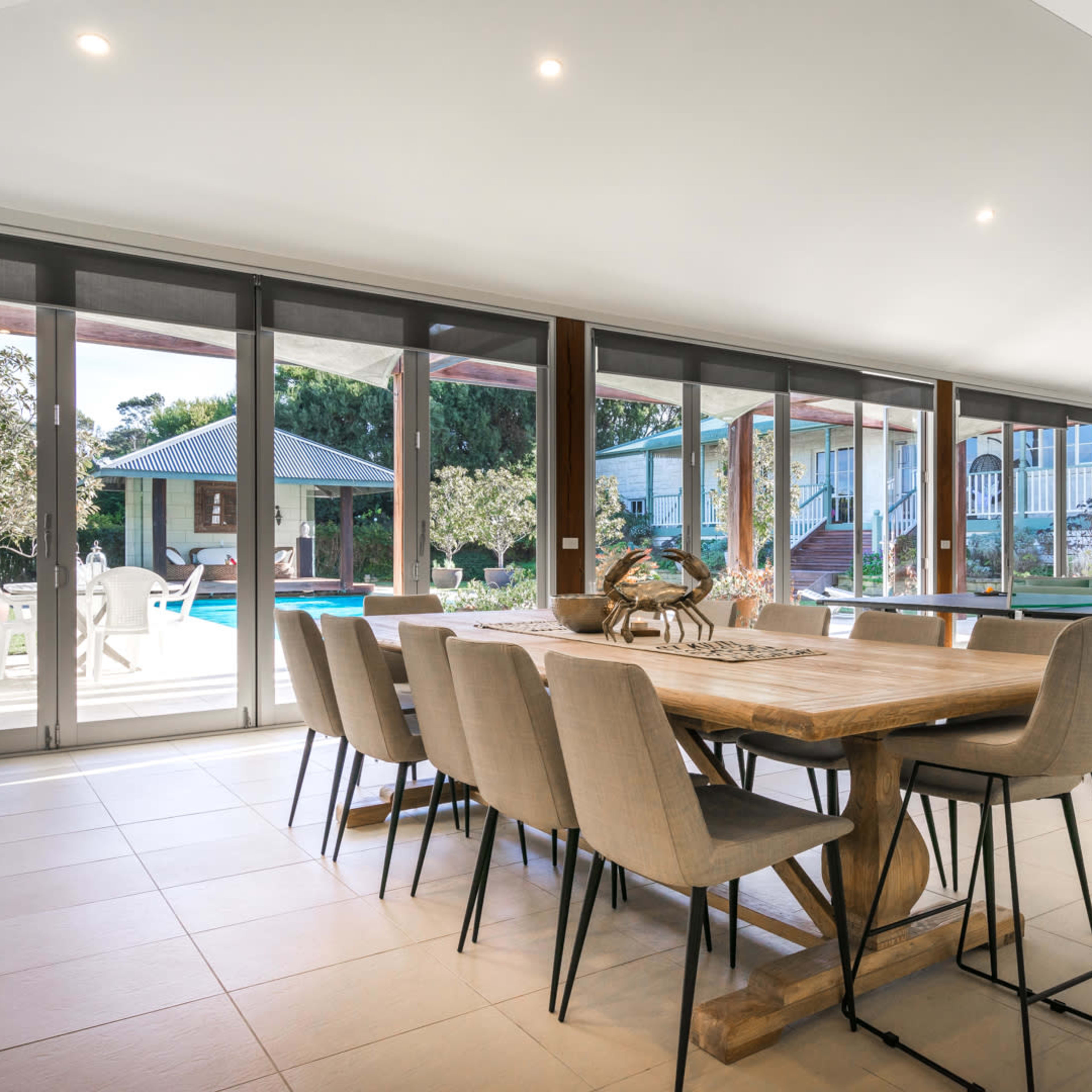 A spacious dining area features a long wooden table with upholstered chairs, large windows overlooking a backyard with a pool, and a modern decor.