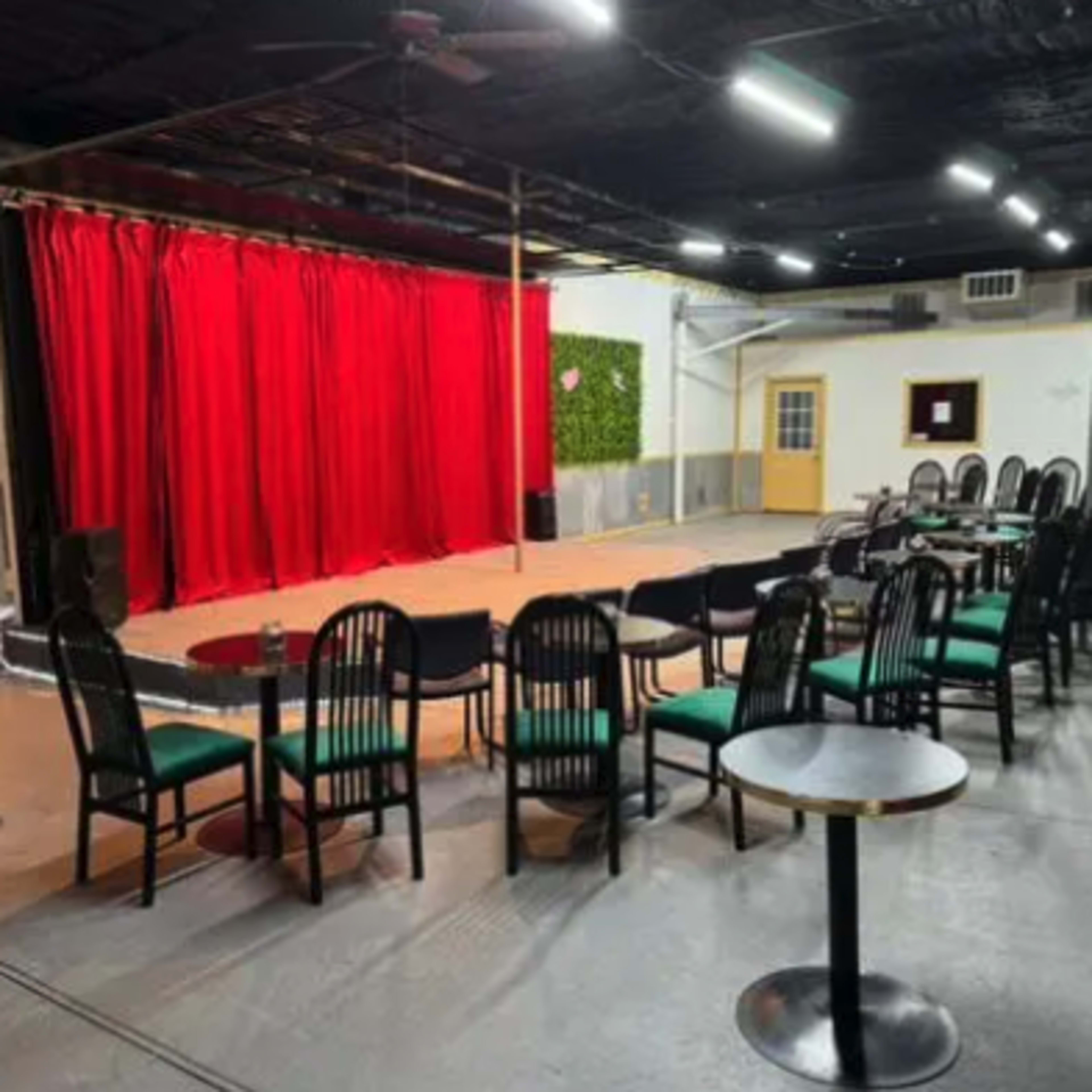 A performance venue with a red curtain, a stage area, and rows of black chairs arranged around small tables.