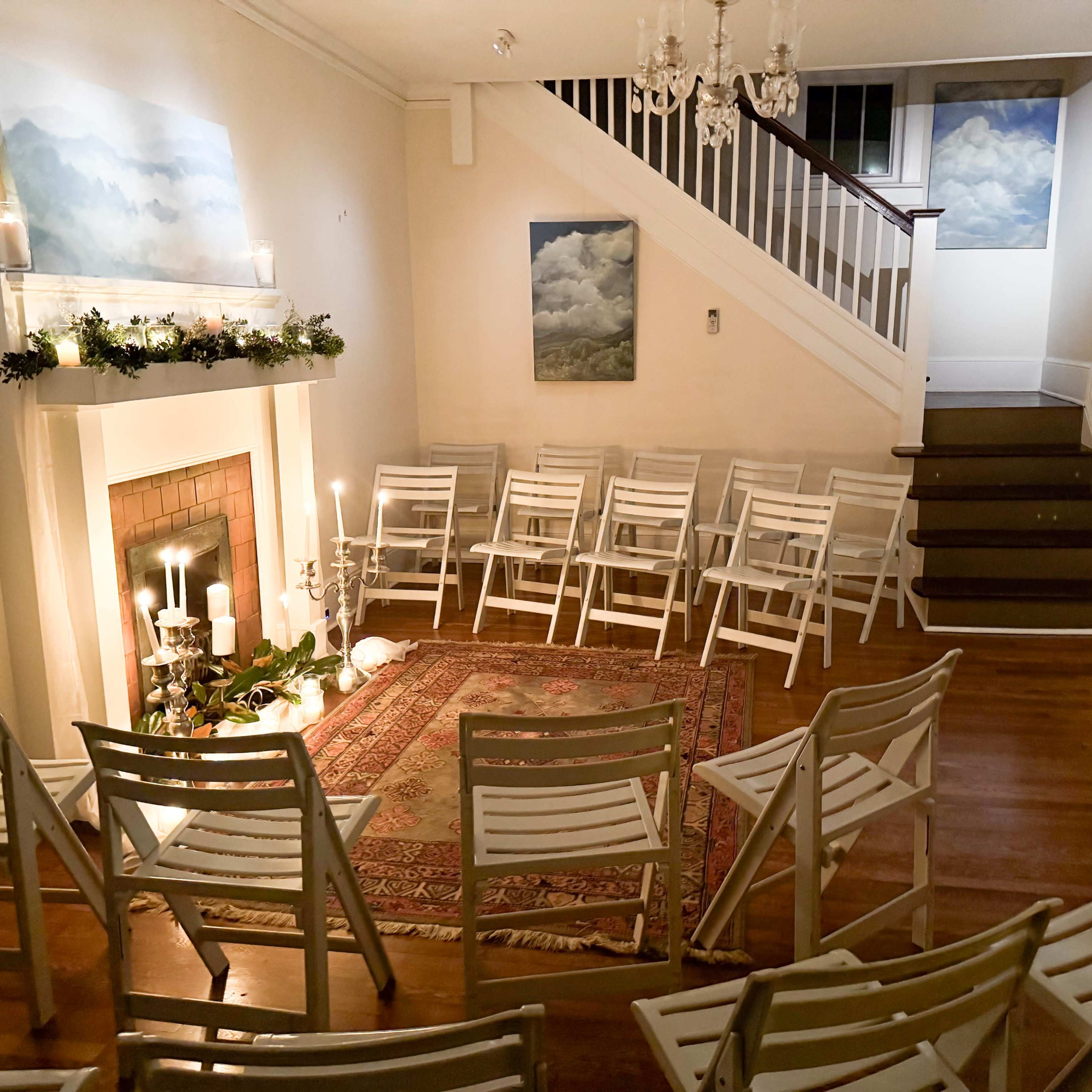 A neatly arranged room with white chairs set up around a decorative area featuring a rug and a fireplace, with paintings of clouds on the walls.