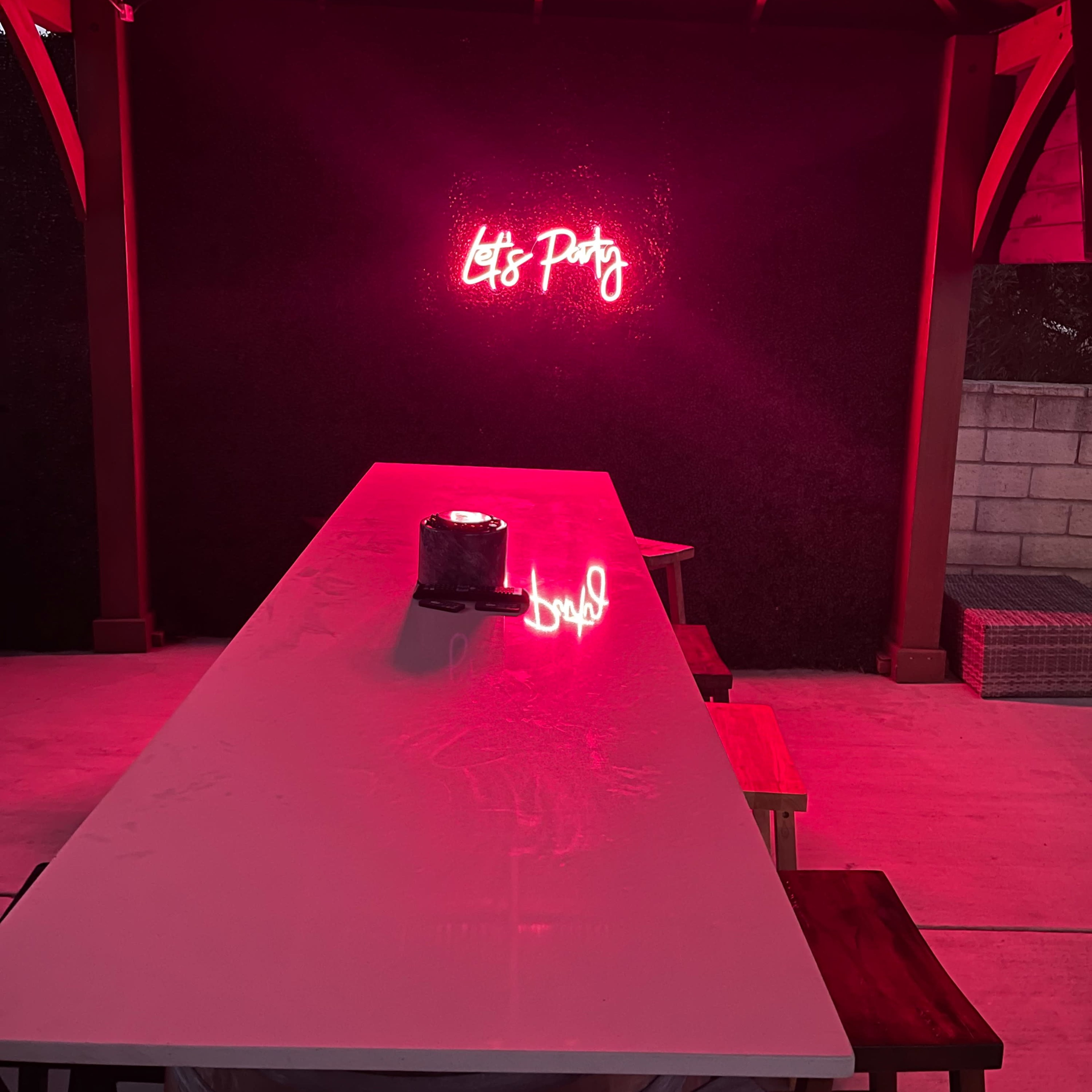A covered outdoor space with a long table, a candle in the center, and a neon sign that reads "Let's Party" on the wall.