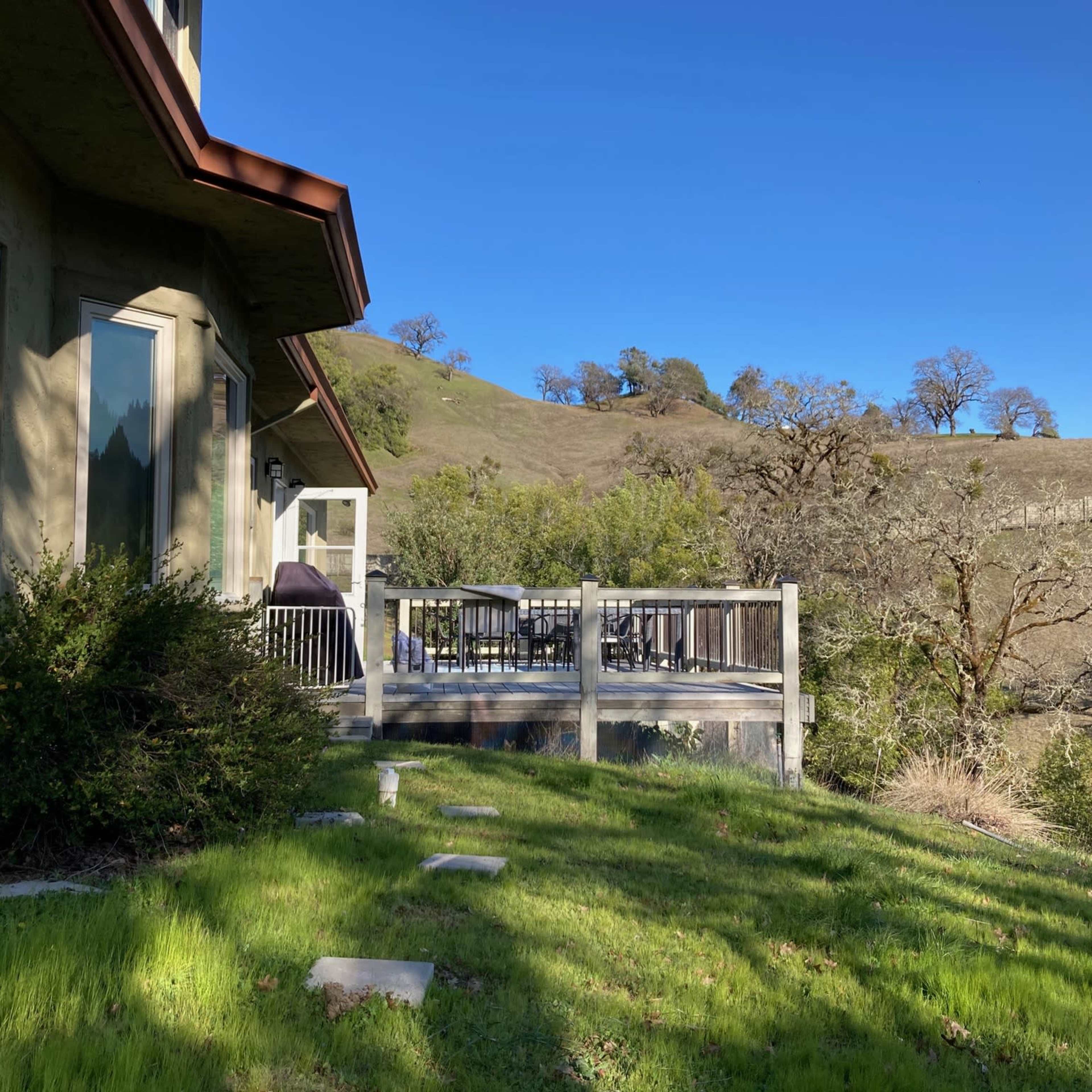 A house with large windows and a red roof is set beside a grassy area and a wooden deck, against a backdrop of rolling hills and trees.