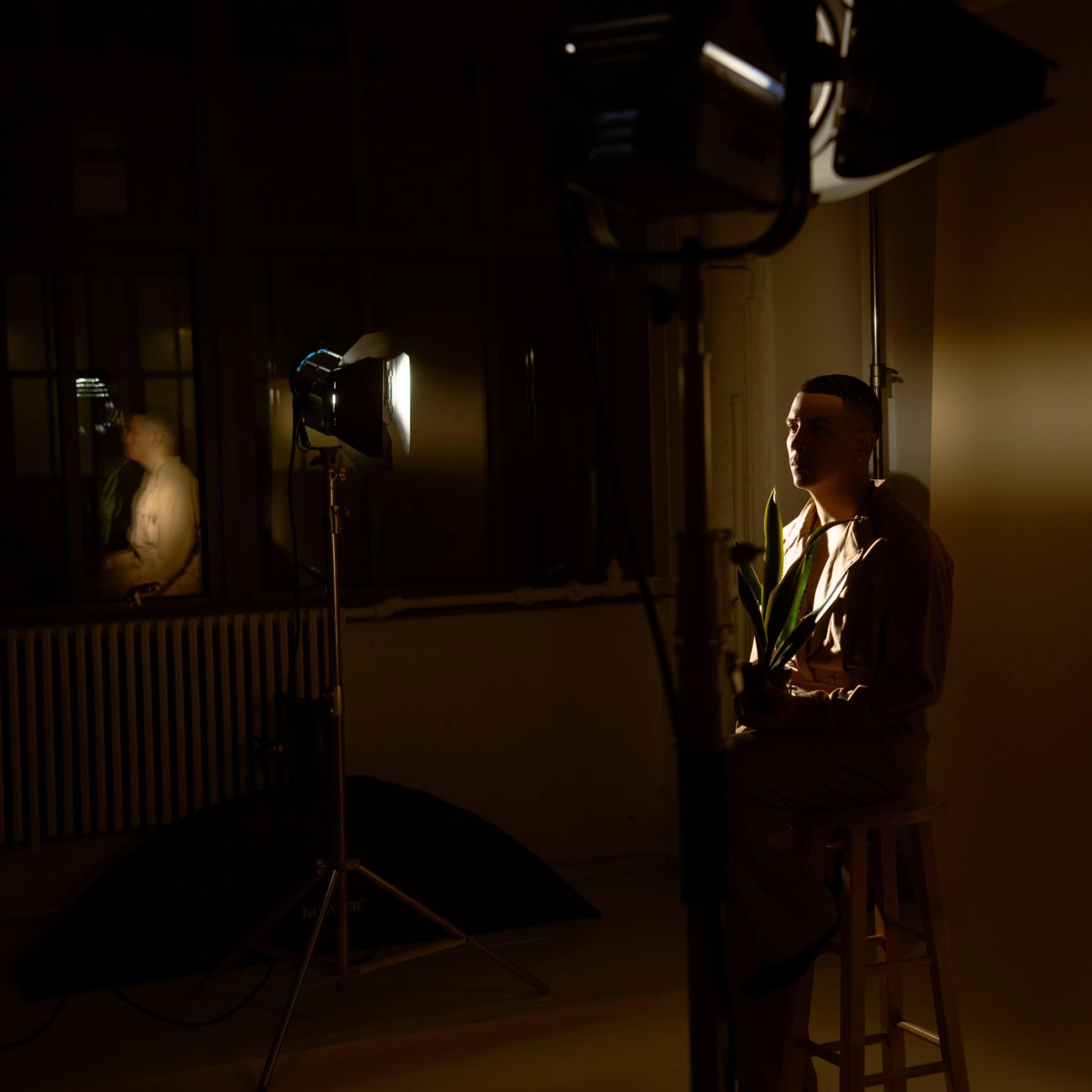 A person sits on a stool in a well-lit studio surrounded by lighting equipment against a blank backdrop.