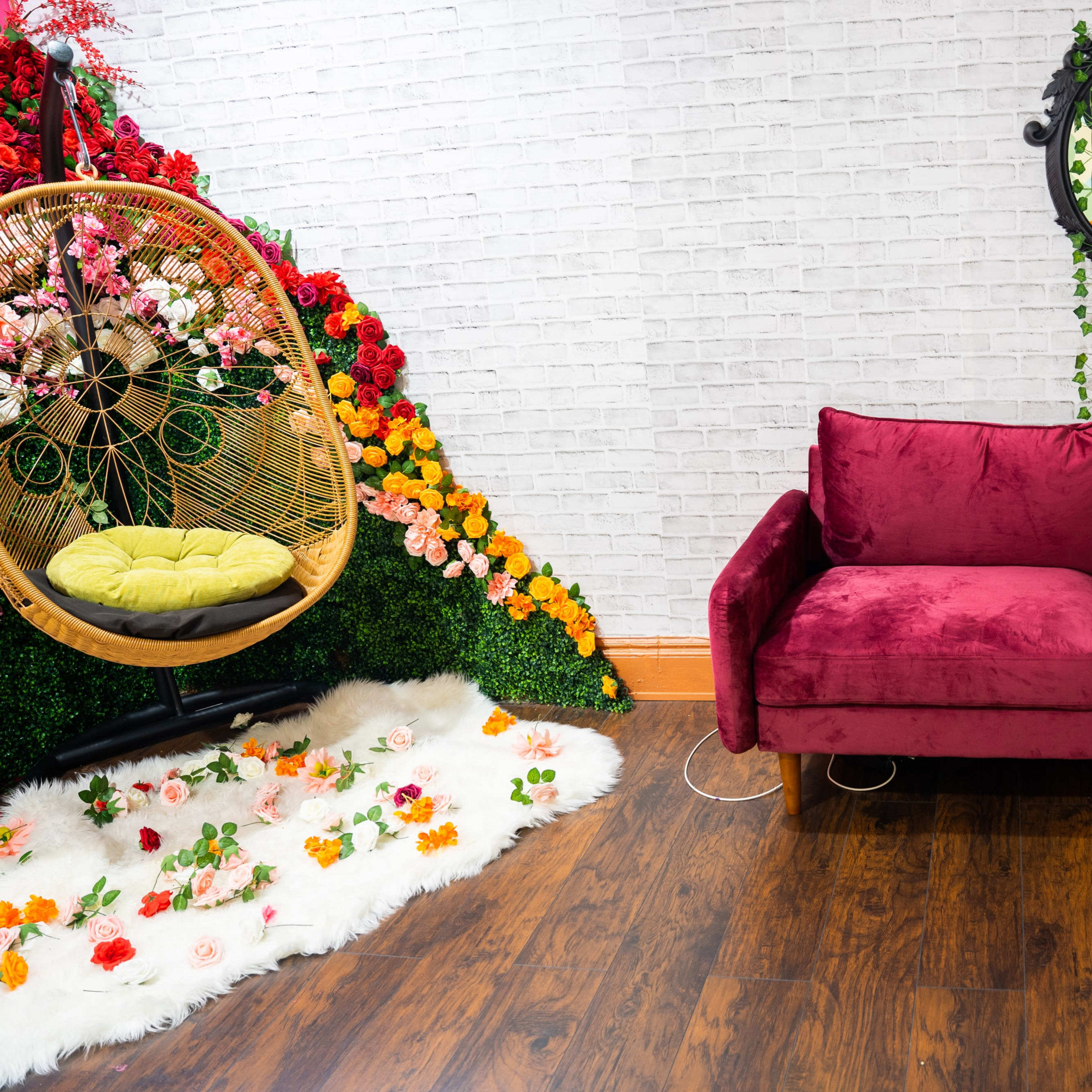 A vibrant room with a maroon sofa, a hanging chair surrounded by colorful flowers, and a mirror adorned with greenery.