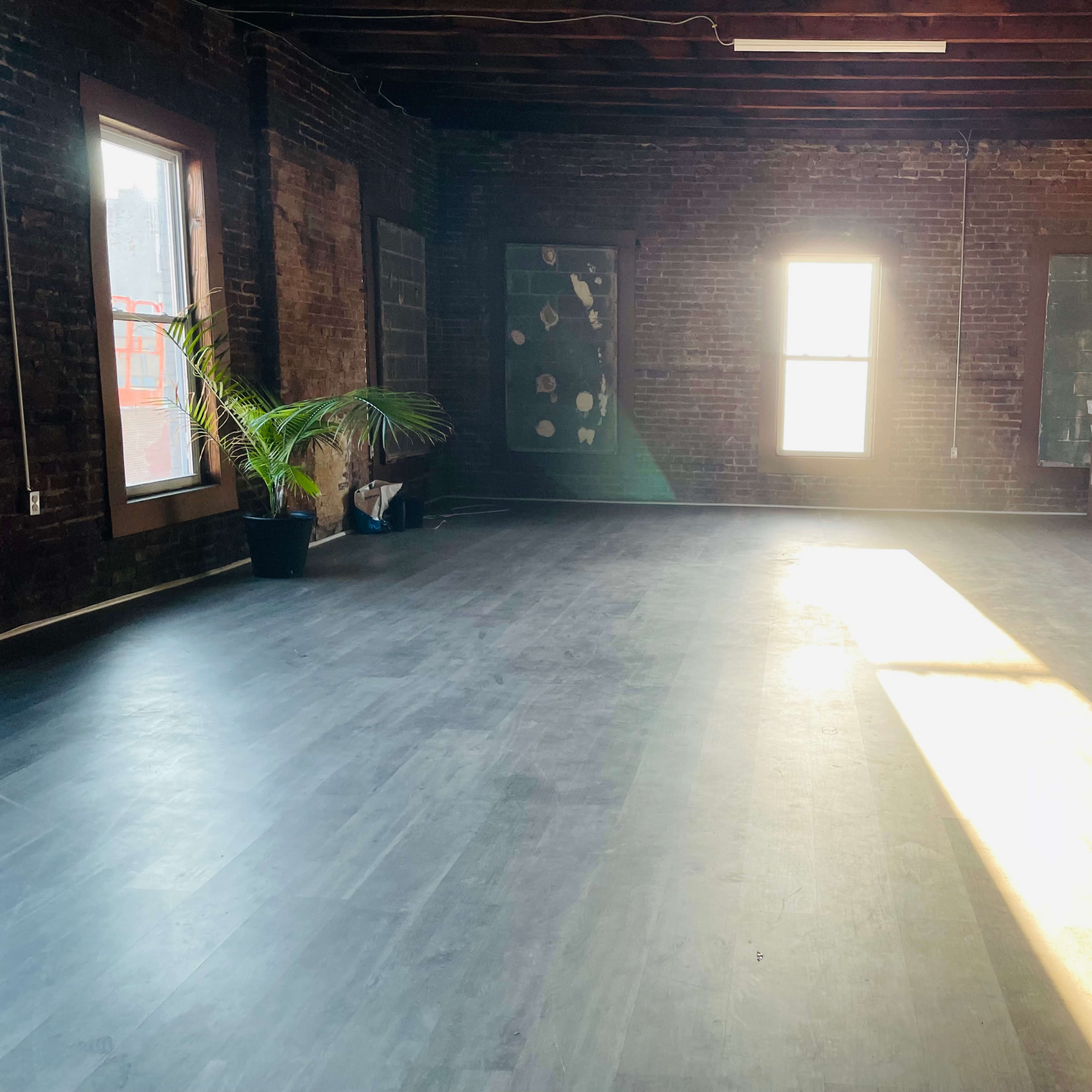 An empty room with exposed brick walls, large windows allowing sunlight to stream in, and a potted plant in one corner.
