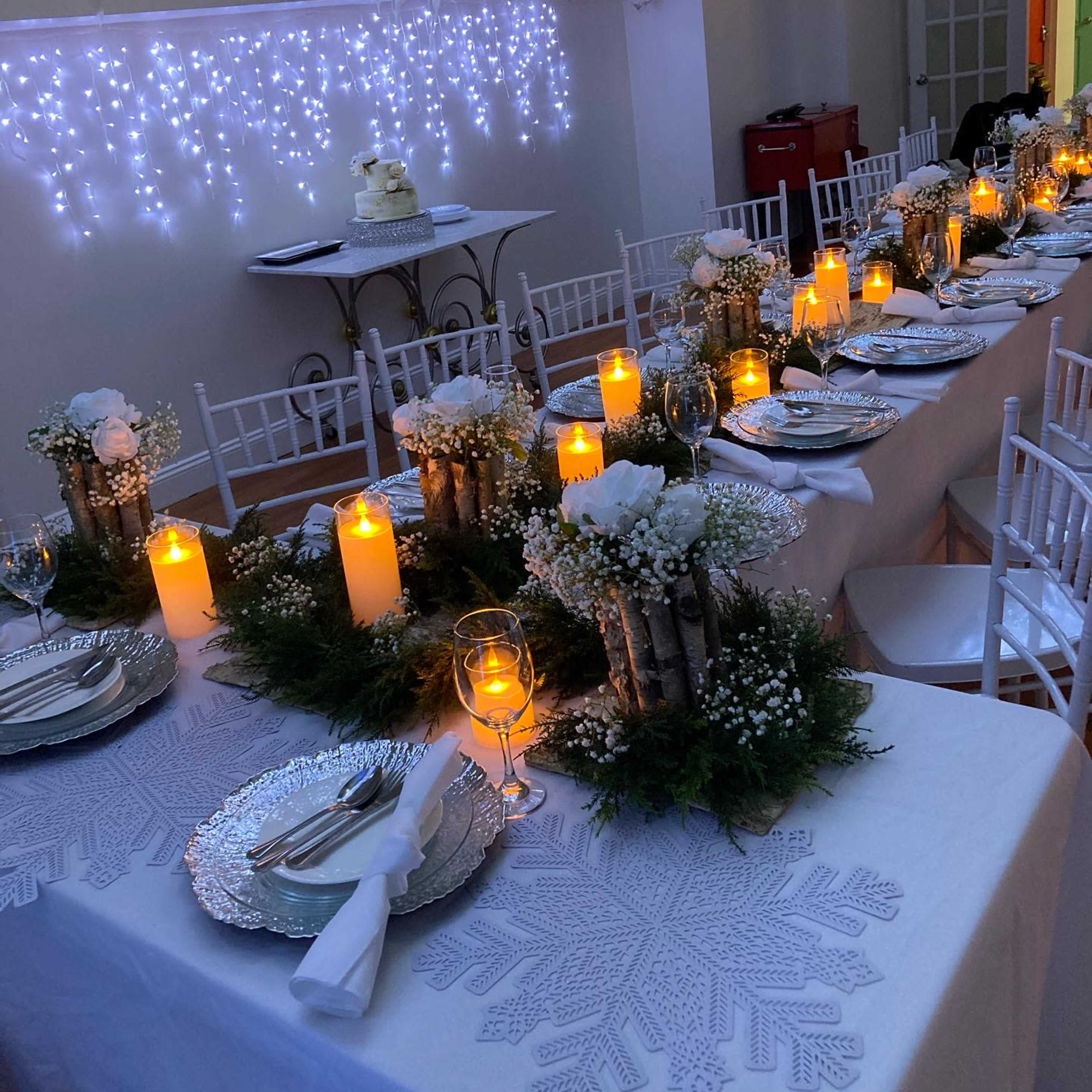 A long dining table is set with white tablecloths, silver plates, candles, and floral arrangements, all under a backdrop of hanging lights and decorations.
