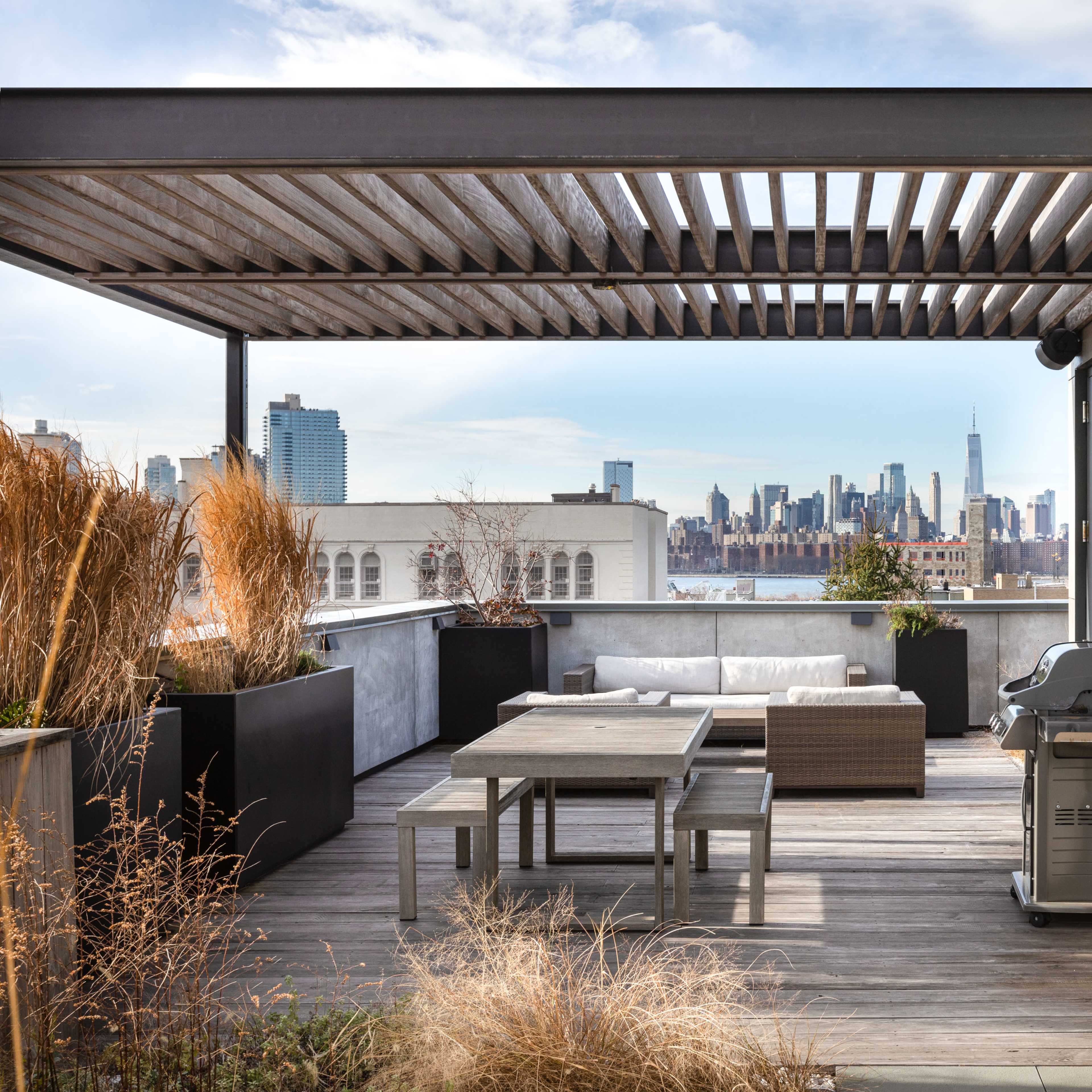 A rooftop terrace featuring a table, seating area, and grill, with a city skyline visible in the background.