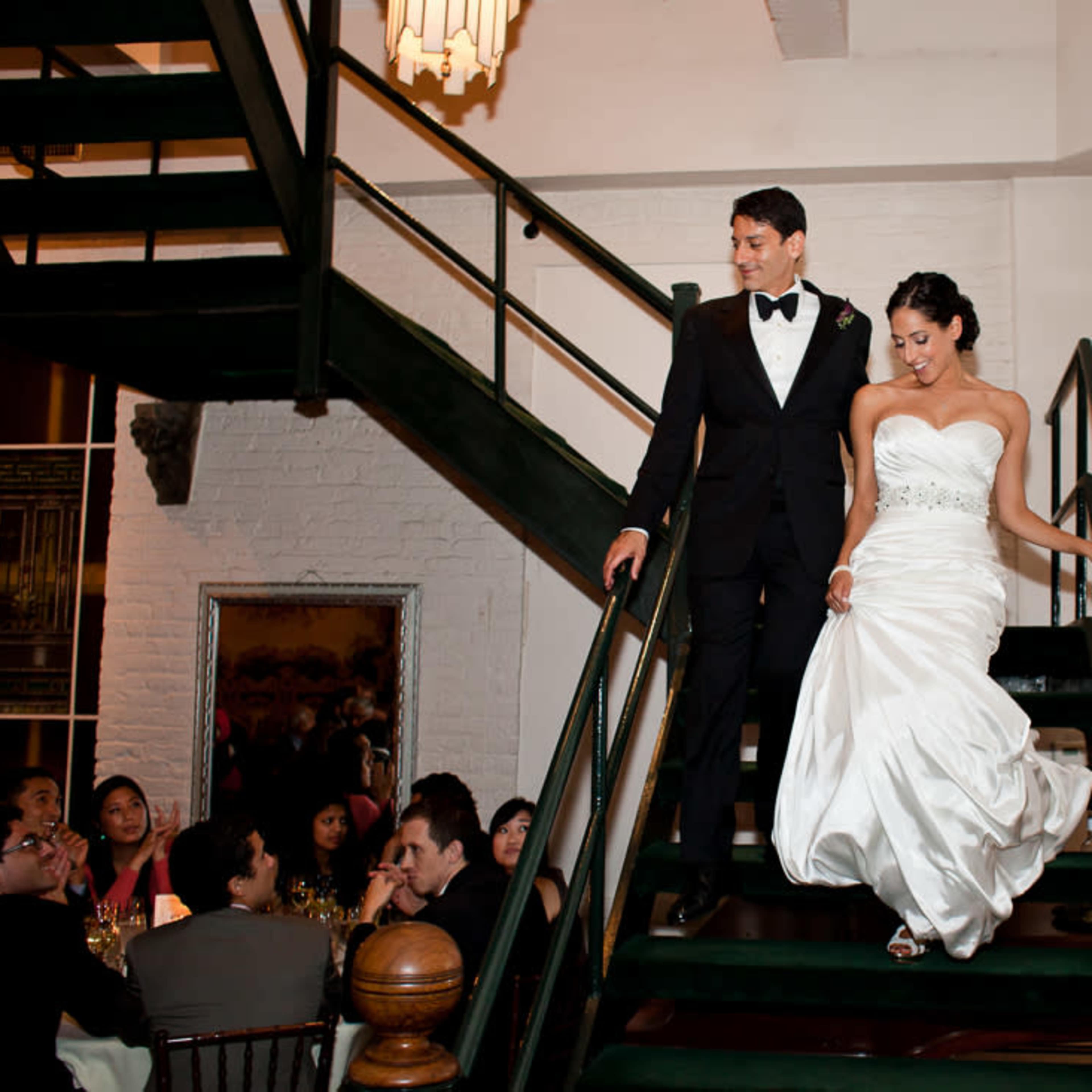A bride and groom descend a staircase in a banquet hall while guests sit at tables below.