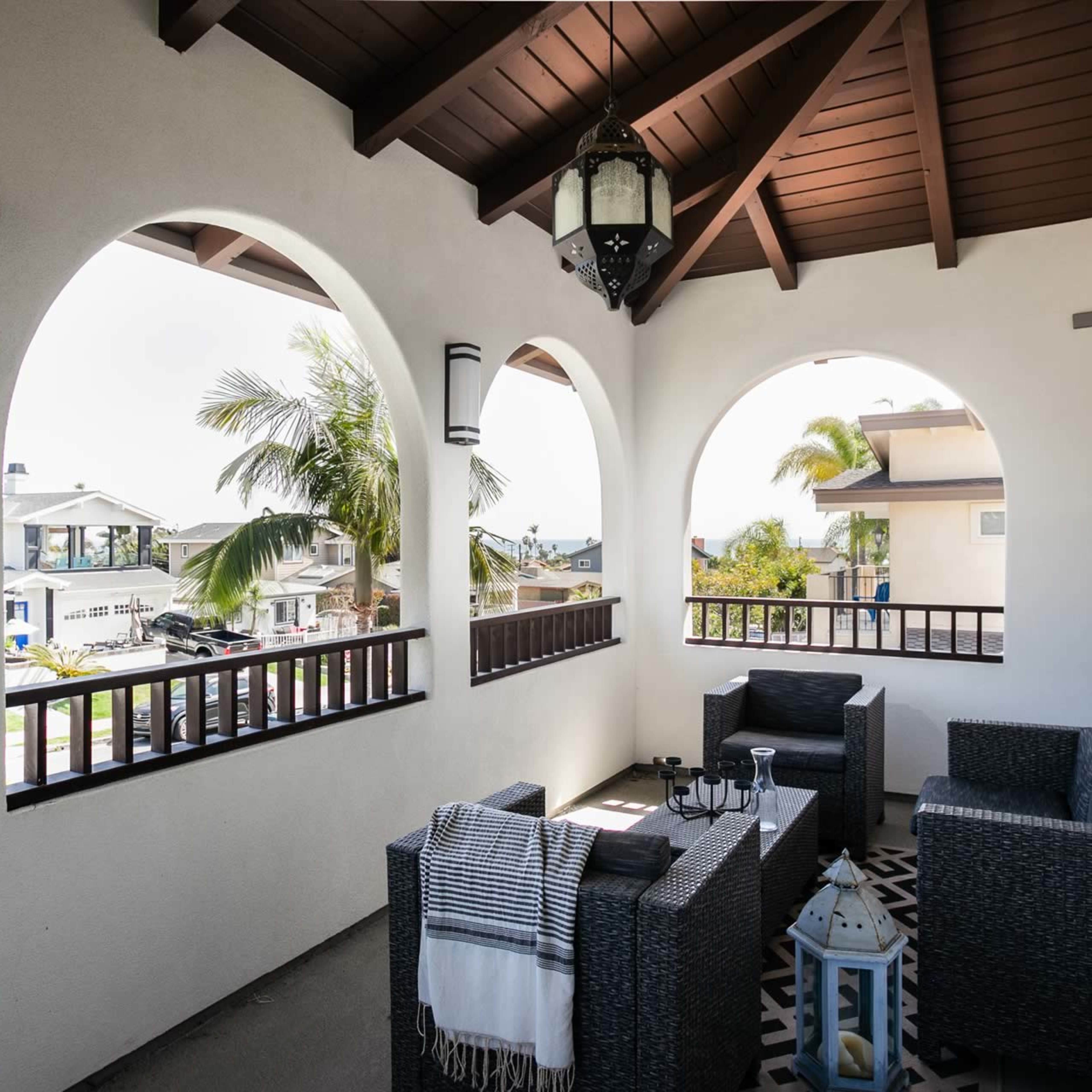 A covered balcony with arched openings, featuring wicker furniture, a lantern, and decorative tiles, overlooking a residential area.