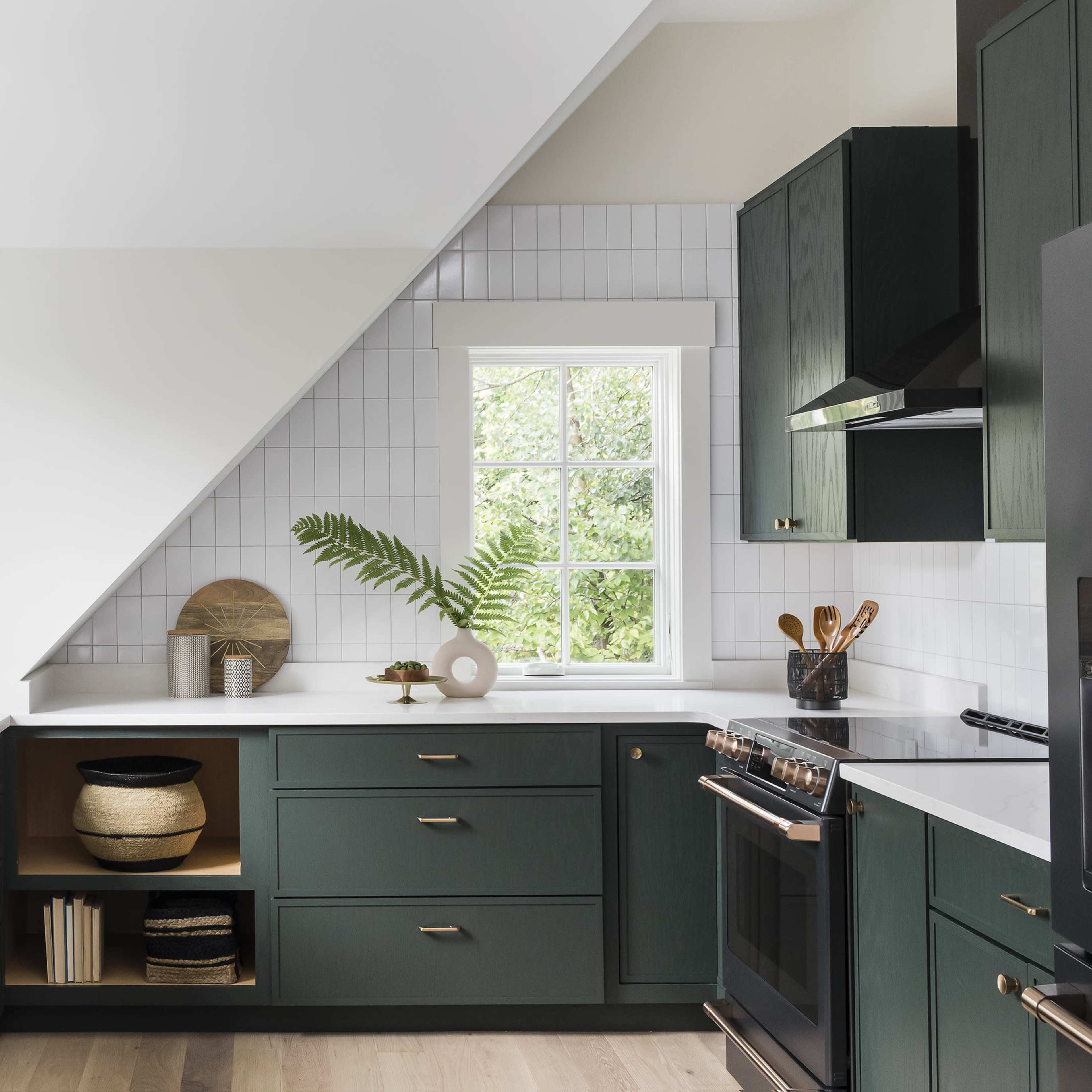 A modern kitchen features dark green cabinetry, white countertops, and a large window with natural light illuminating the space.