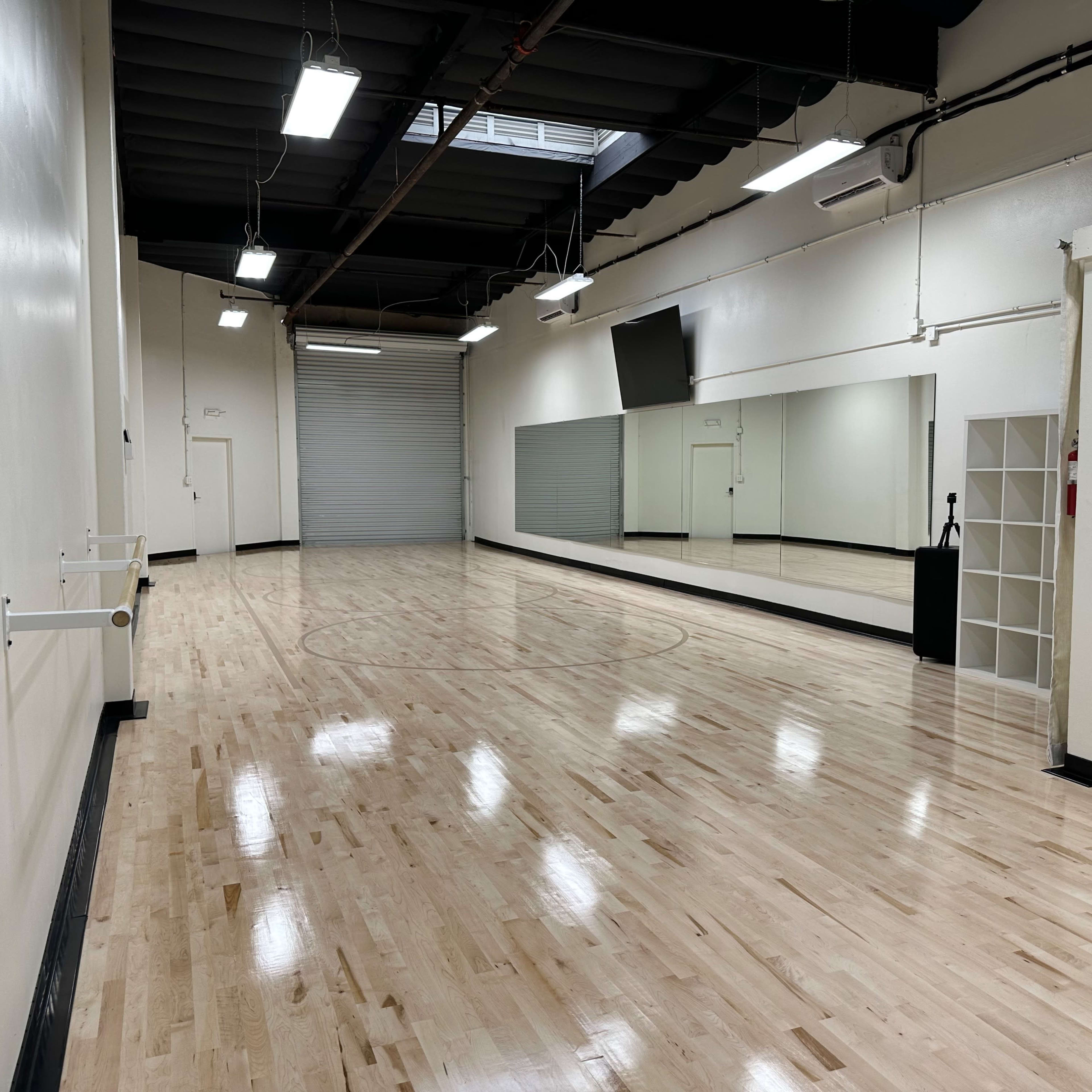 A spacious, empty dance studio with polished wooden floors and a large mirror along one wall.