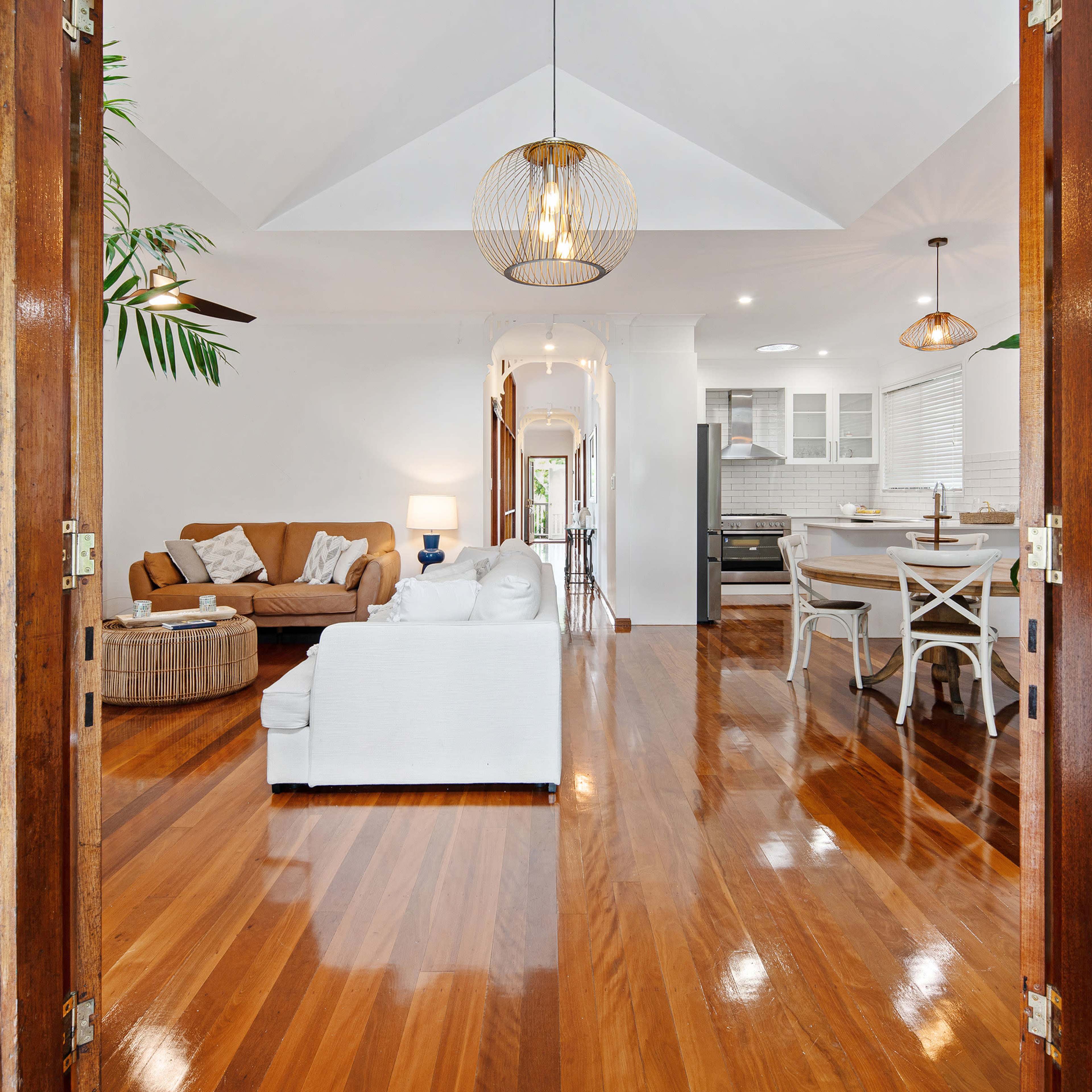 A living room features wooden flooring, a white sofa, brown chairs, and large plants, with light streaming through open double doors.