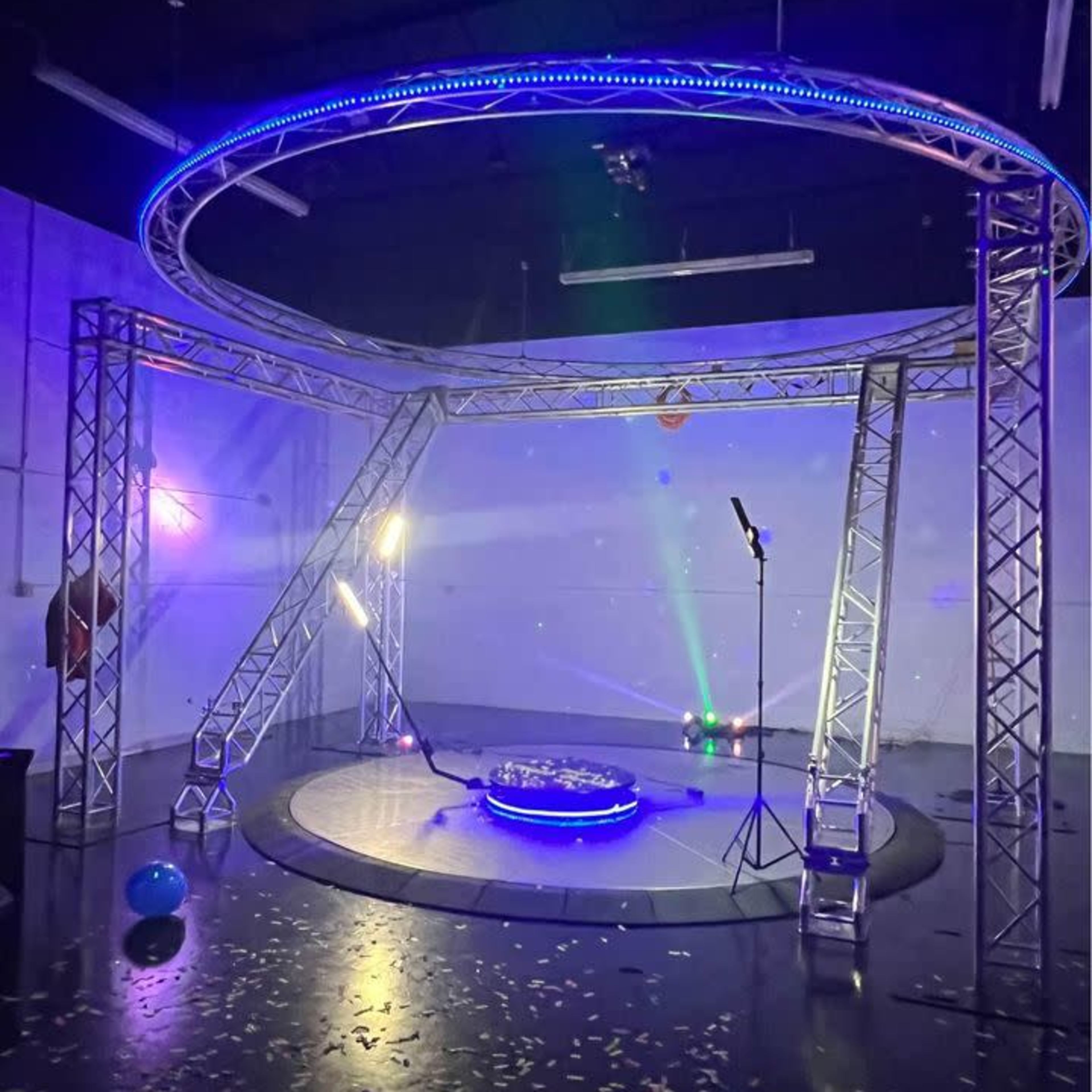 A circular stage structure with a lighting setup and reflective surface, surrounded by a sparse, decorated room with confetti on the floor.