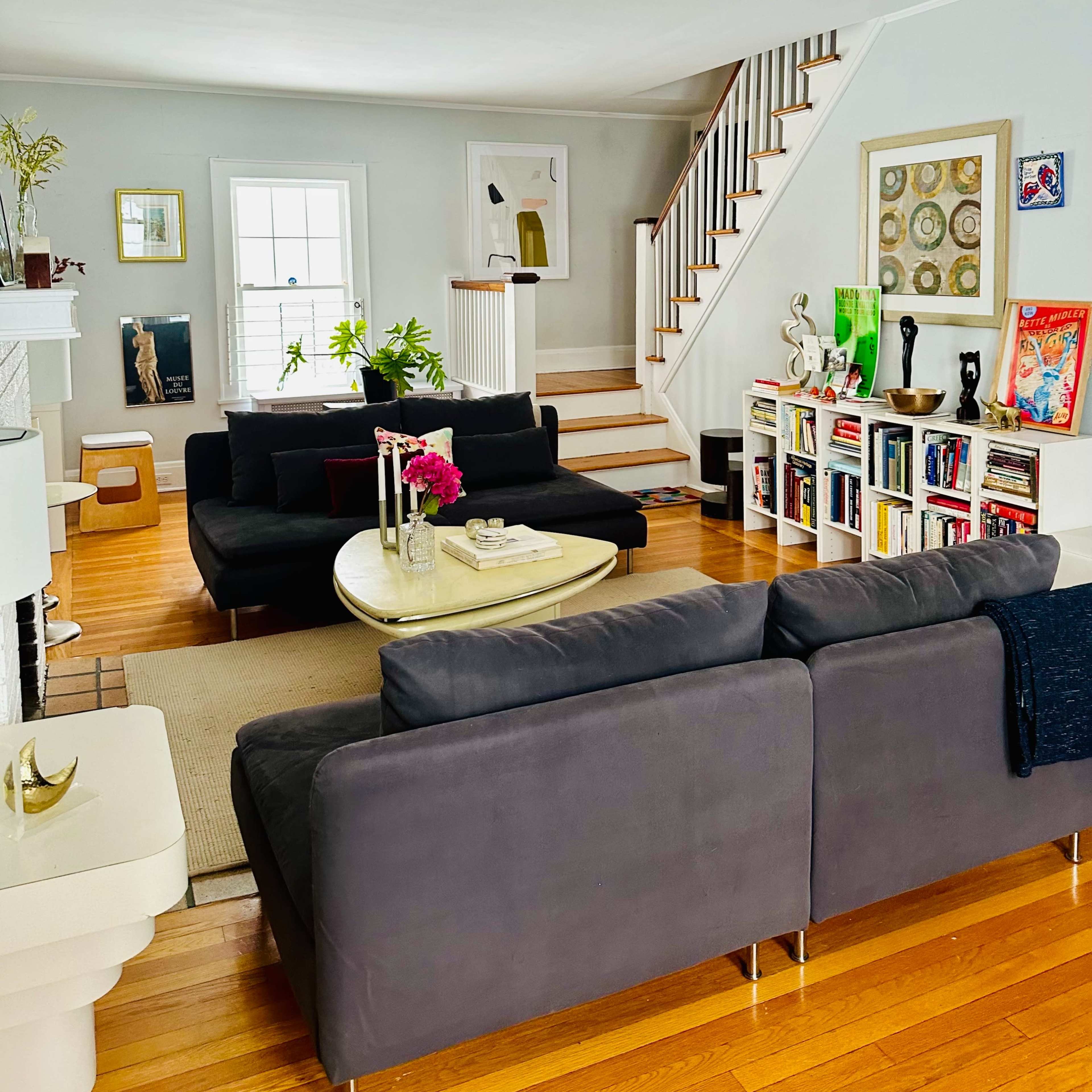 The living room features two dark sofas facing a central coffee table, with a staircase and a bookshelf in the background.
