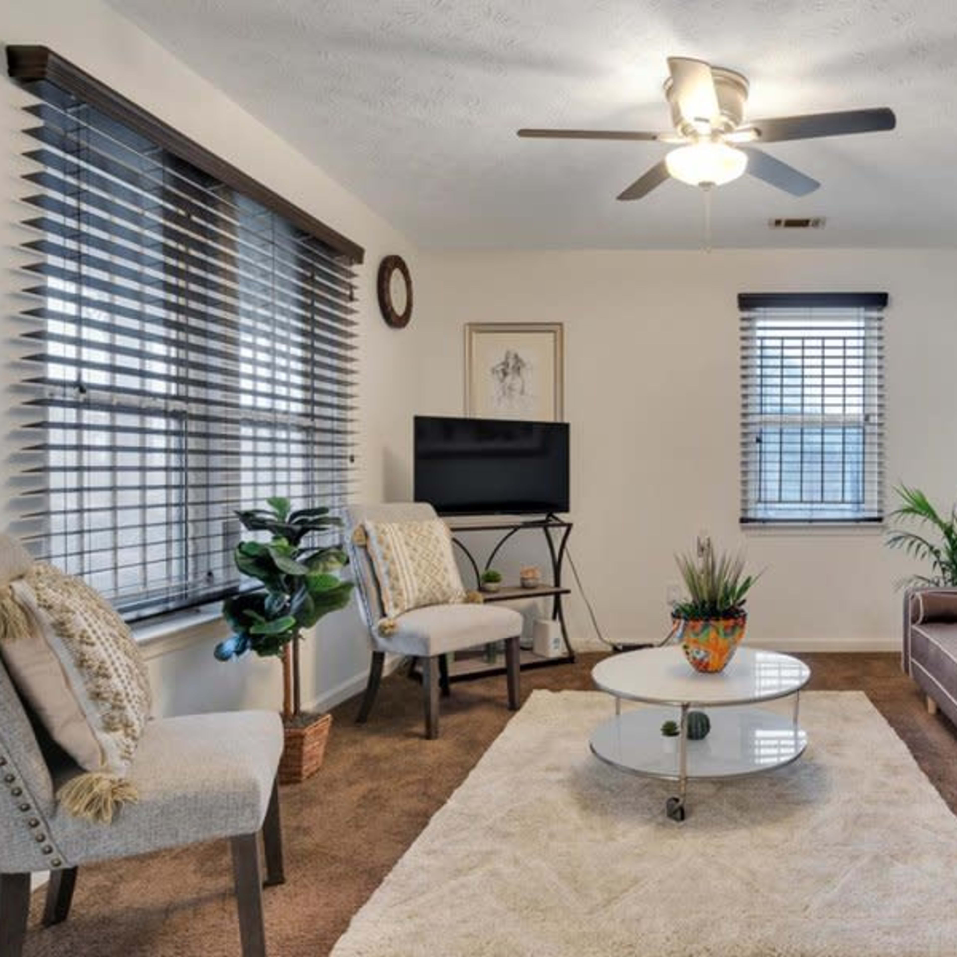 The living room features a sofa, two chairs, a round coffee table, and a television, all arranged around a carpeted area with large windows and indoor plants.