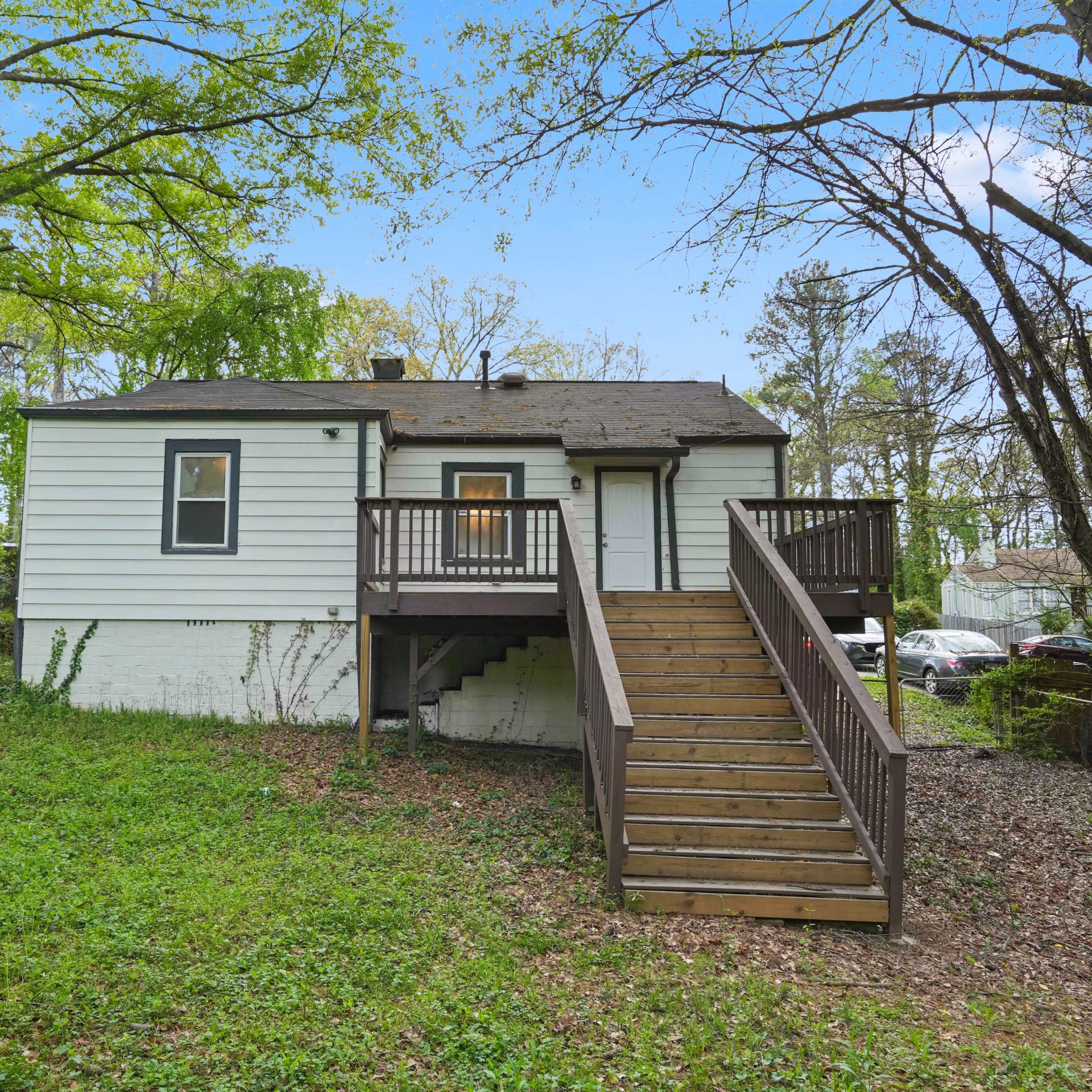 A small, single-story house with a wooden ramp and steps is surrounded by trees and greenery.