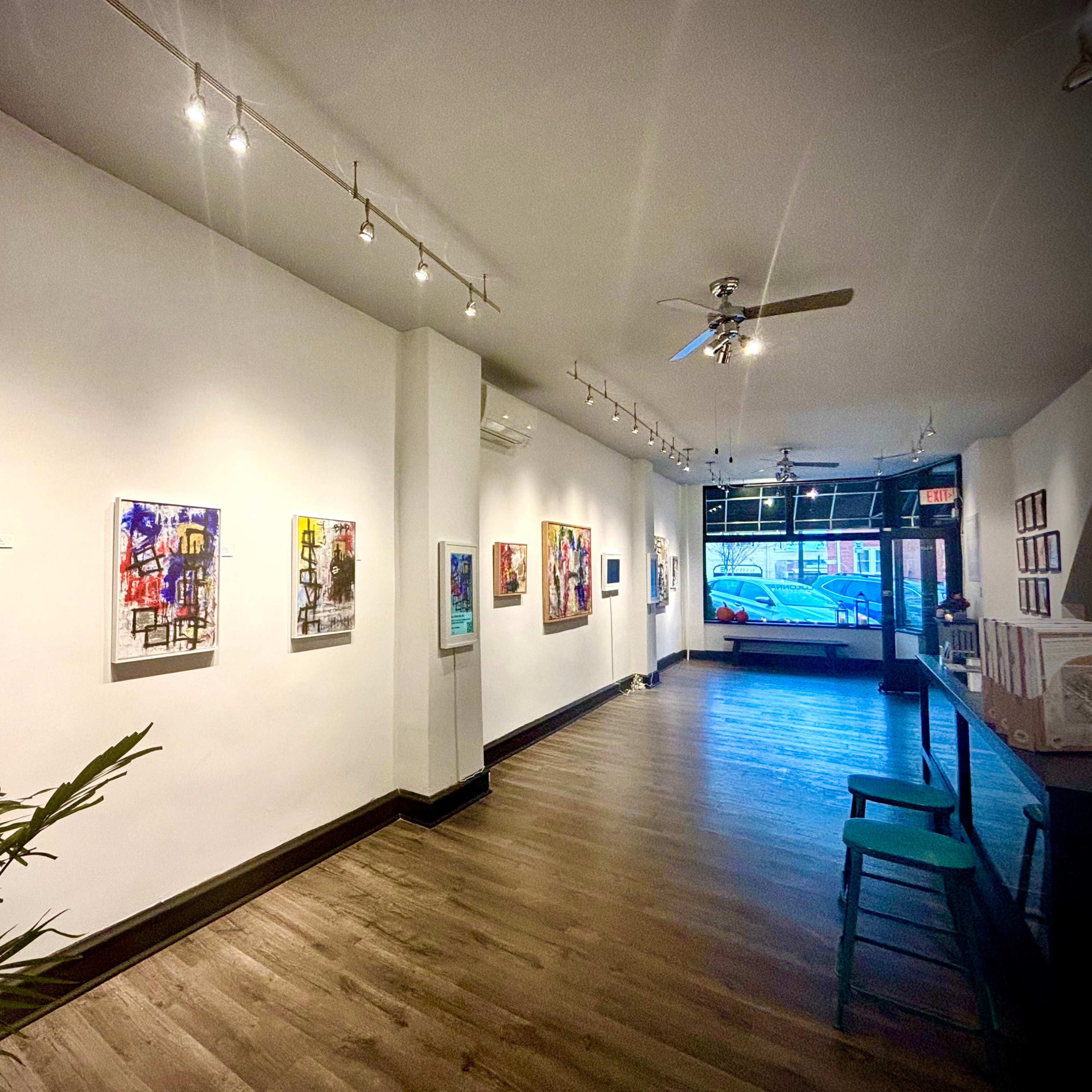 A gallery interior with artwork displayed on white walls and wooden flooring, featuring a row of paintings on one side and a seating area on the left.