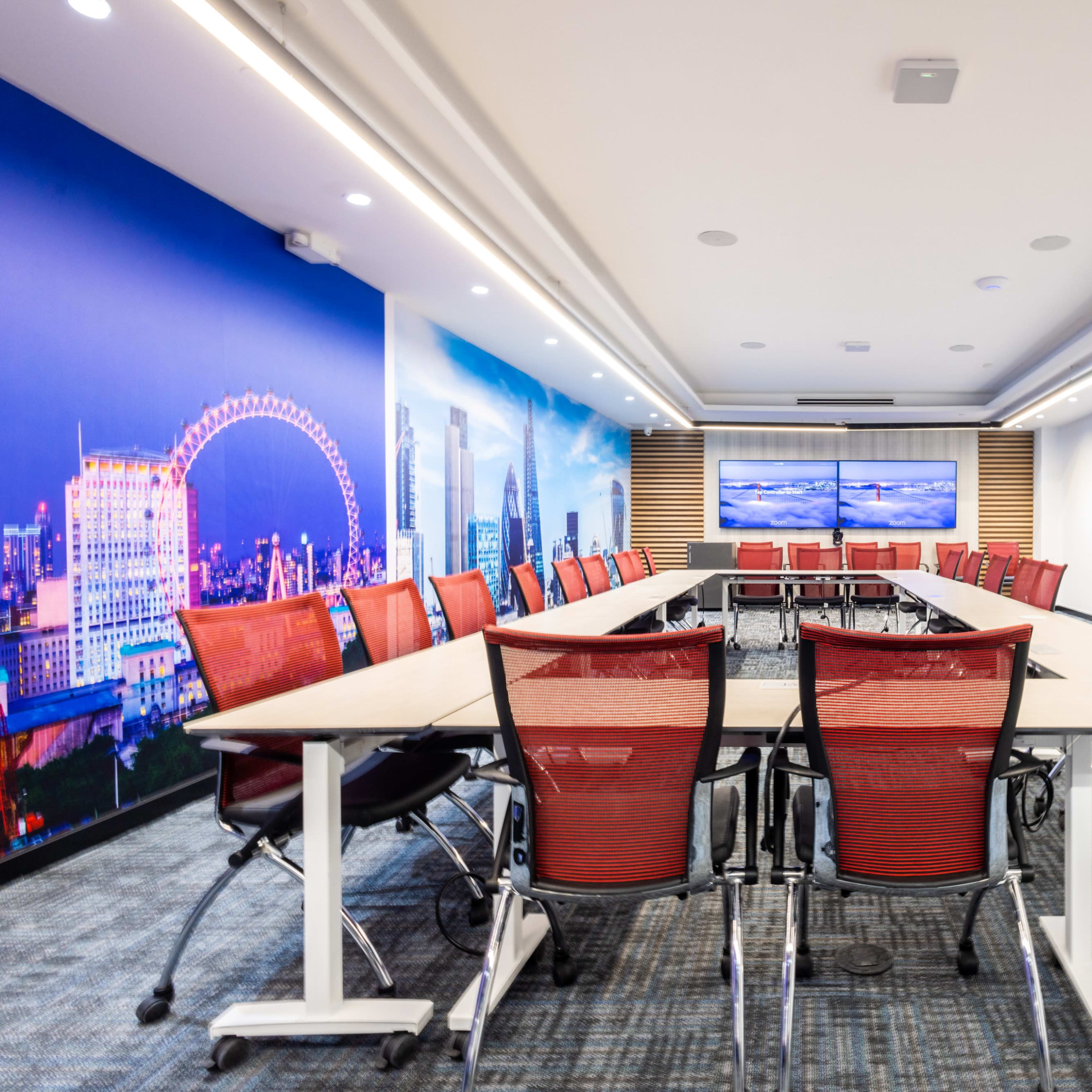A modern conference room with a large table surrounded by red chairs and a backdrop featuring a cityscape.