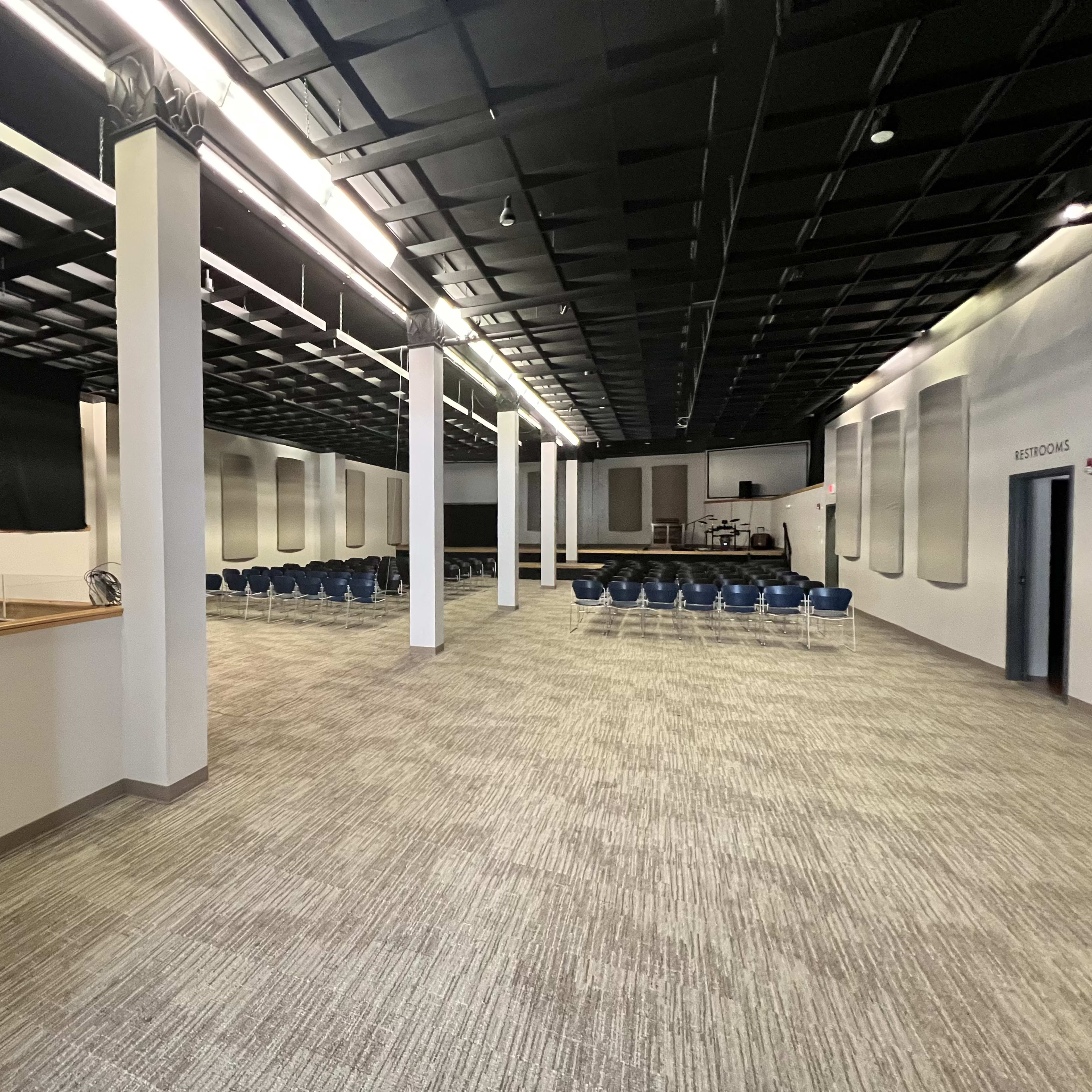 A spacious indoor room with neatly arranged seating, gray walls, and a high ceiling, featuring a stage or platform area at one end.