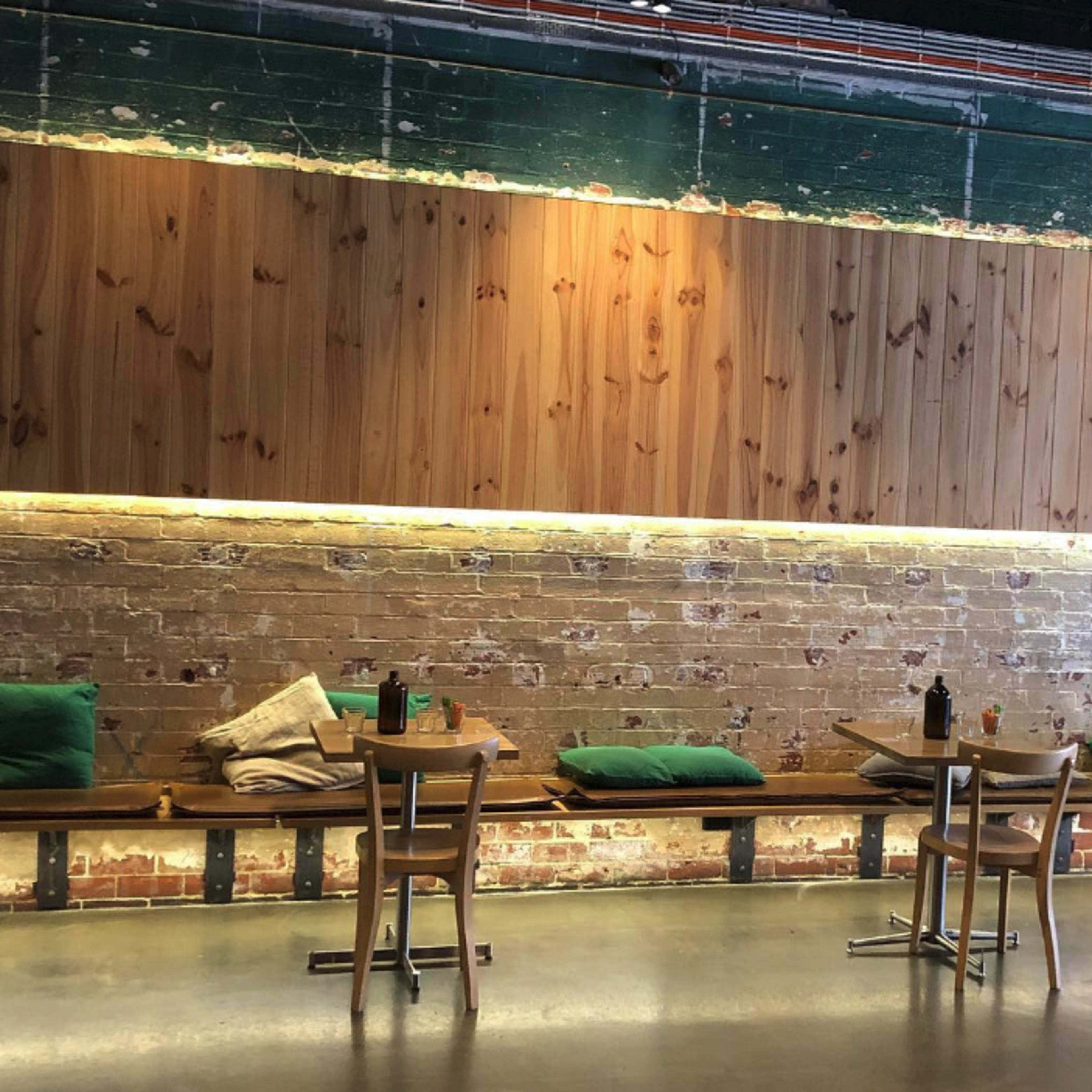 A rustic restaurant interior with wooden walls, a brick backdrop, and wooden tables equipped with green cushions and condiments.