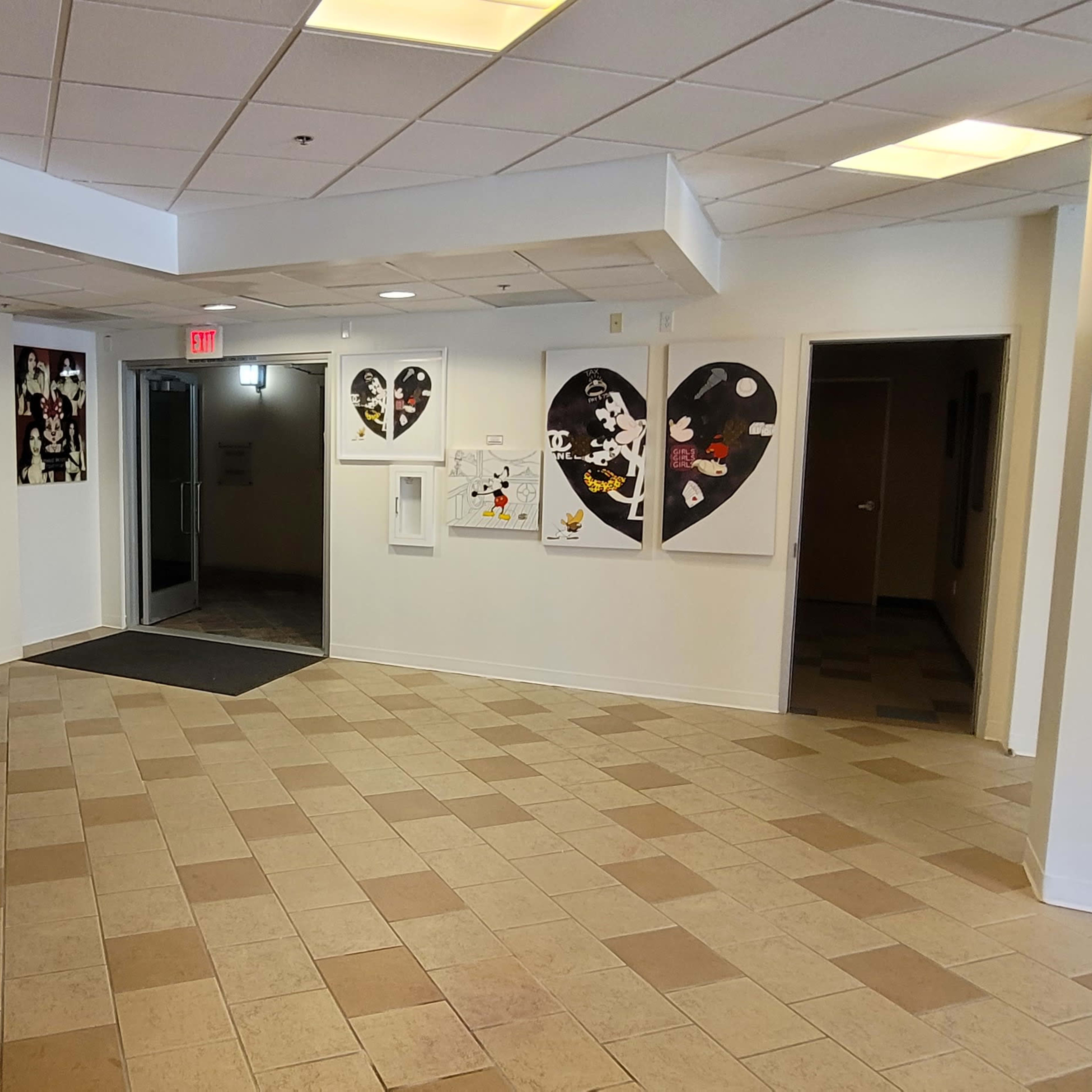 A spacious lobby area with tiled flooring, featuring a reception area, a series of artworks on the walls, and a large television mounted on one wall.