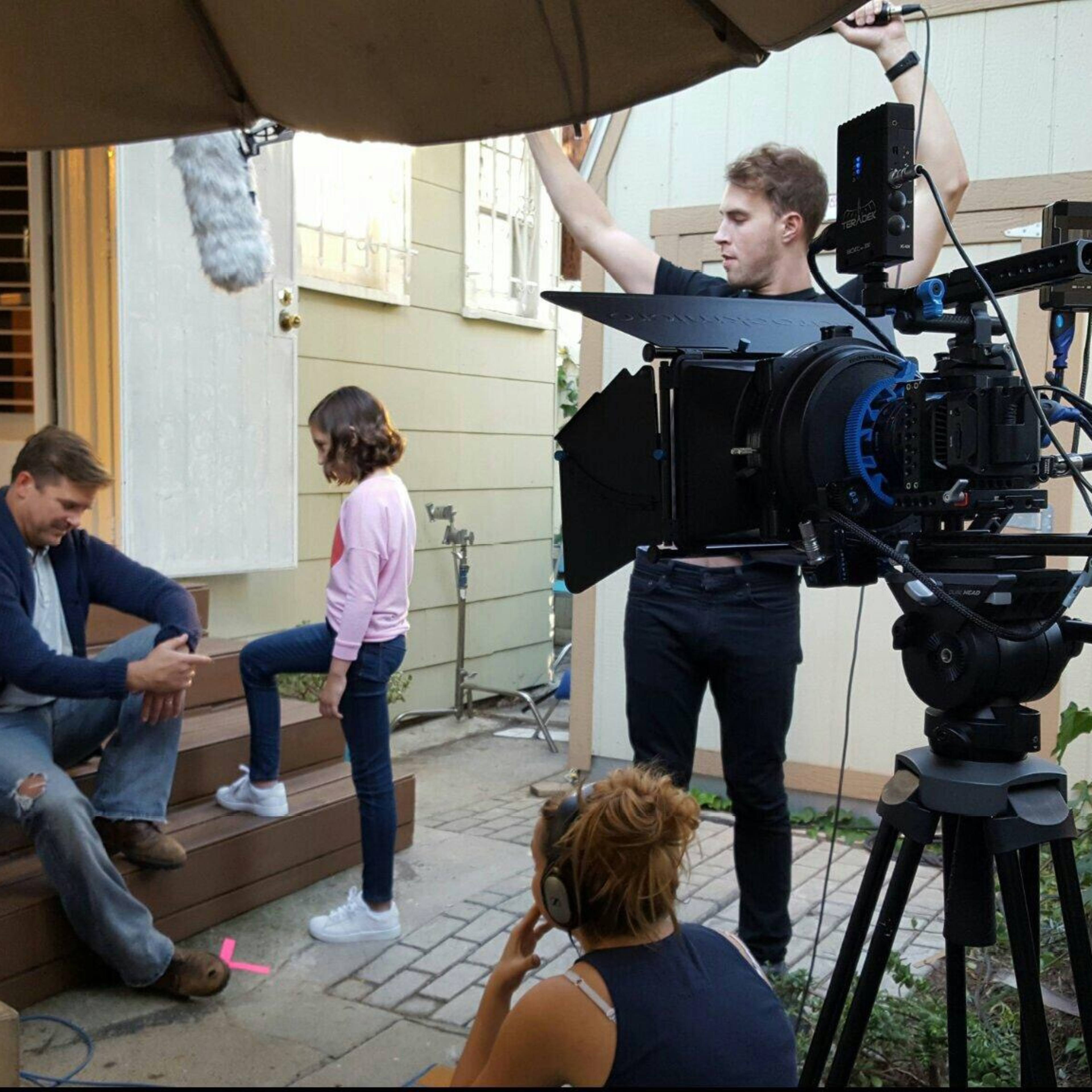 A film crew is setting up a scene on a porch with a man sitting on the steps and a girl standing beside him.