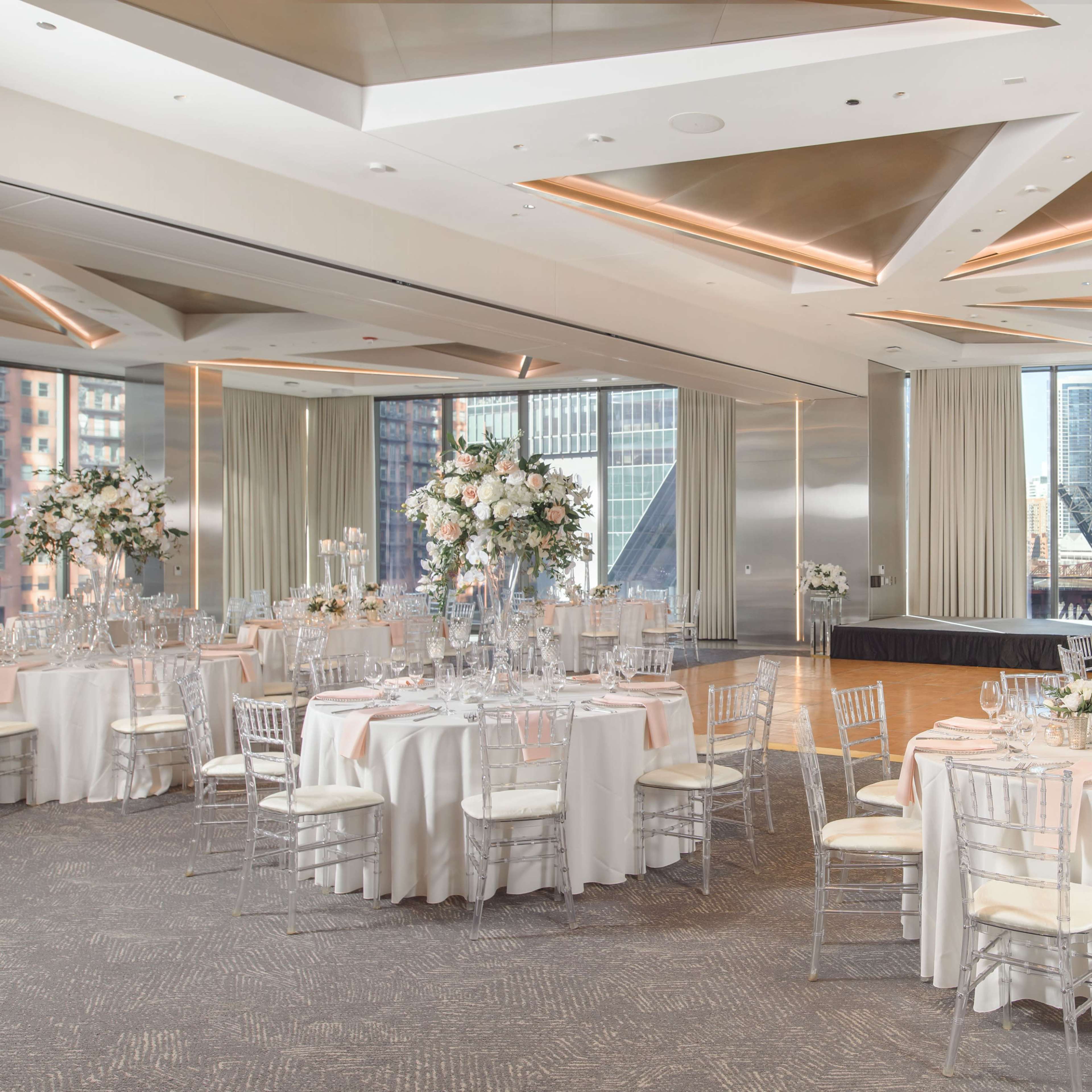 The venue features elegantly arranged round tables with floral centerpieces and a stage, surrounded by large windows showcasing a cityscape.