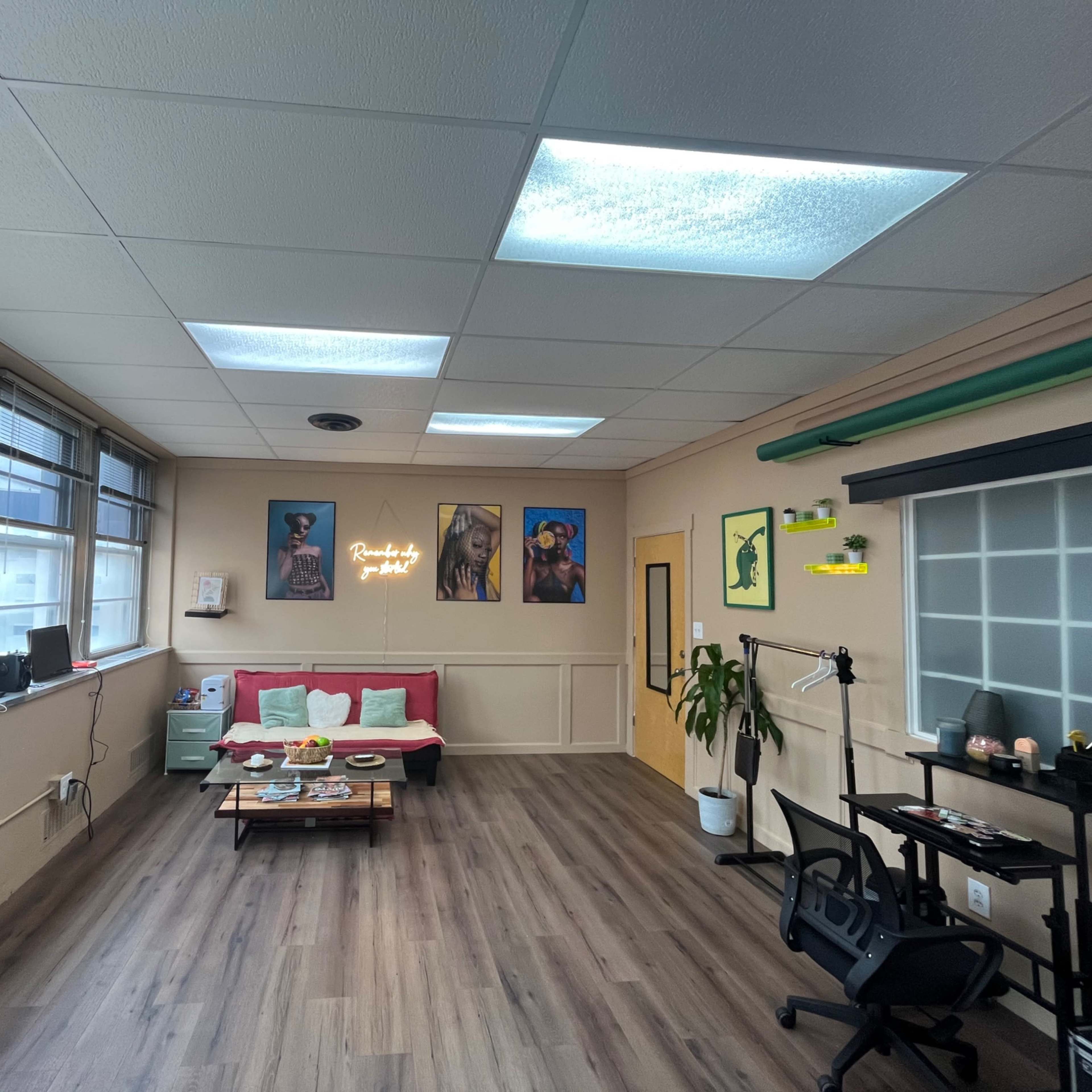 A well-lit office space with a red couch, a coffee table, a desk setup, wall art featuring portraits, and a small plant.
