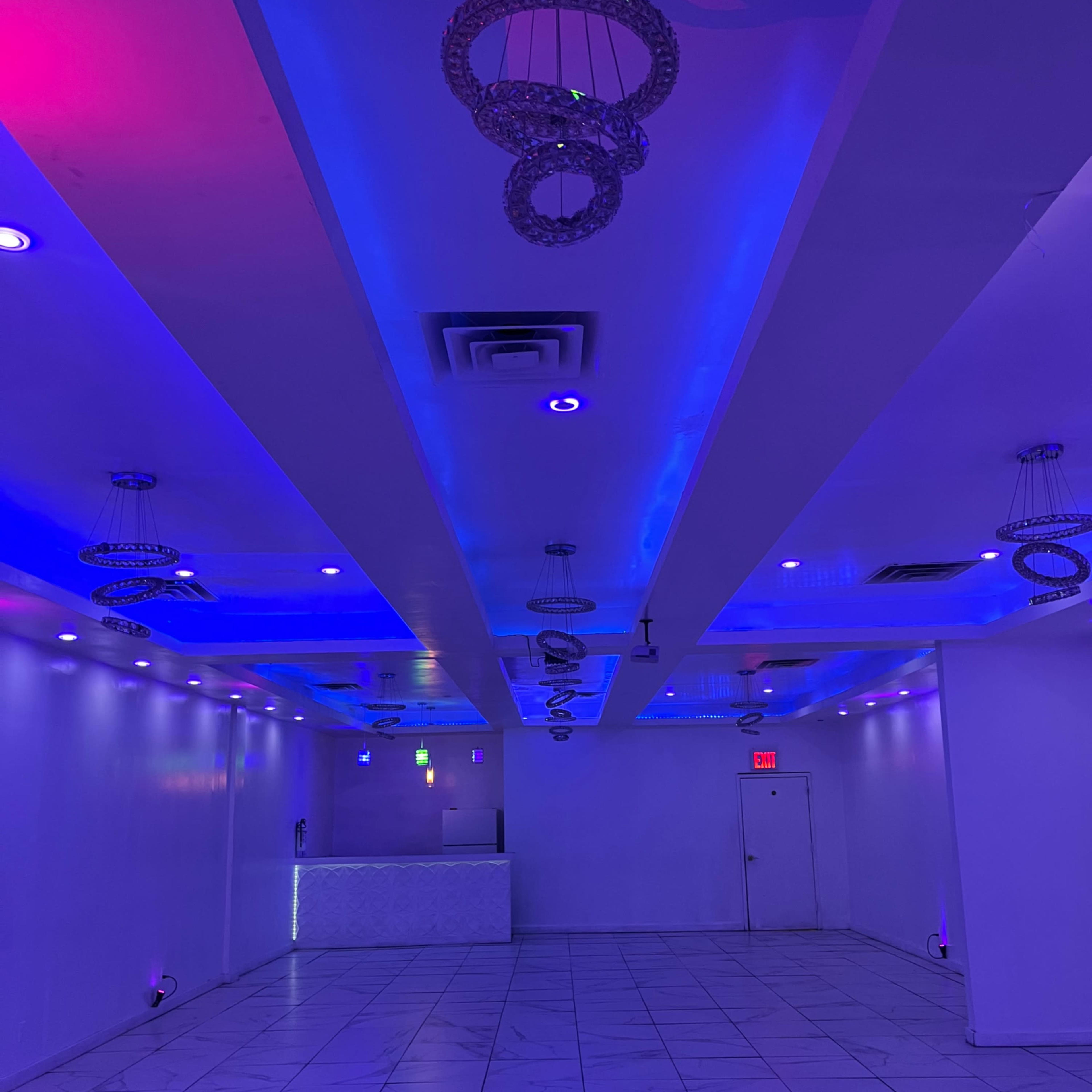 A brightly lit indoor space with a glossy tiled floor, purple and blue lighting, and decorative chandeliers hanging from the ceiling.