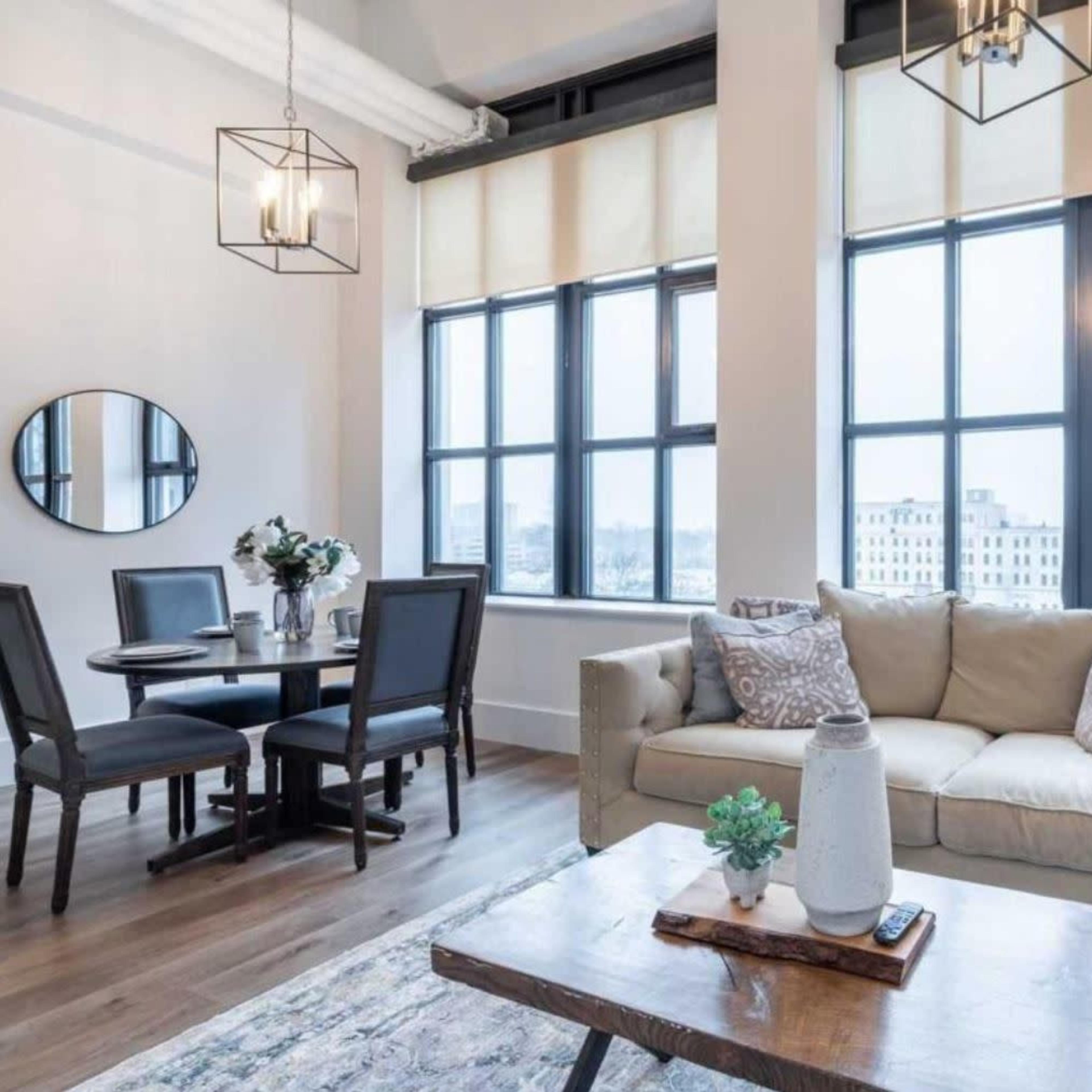 A modern living area featuring a beige sofa, a wooden coffee table, a round dining table with chairs, and large windows allowing natural light to fill the space.