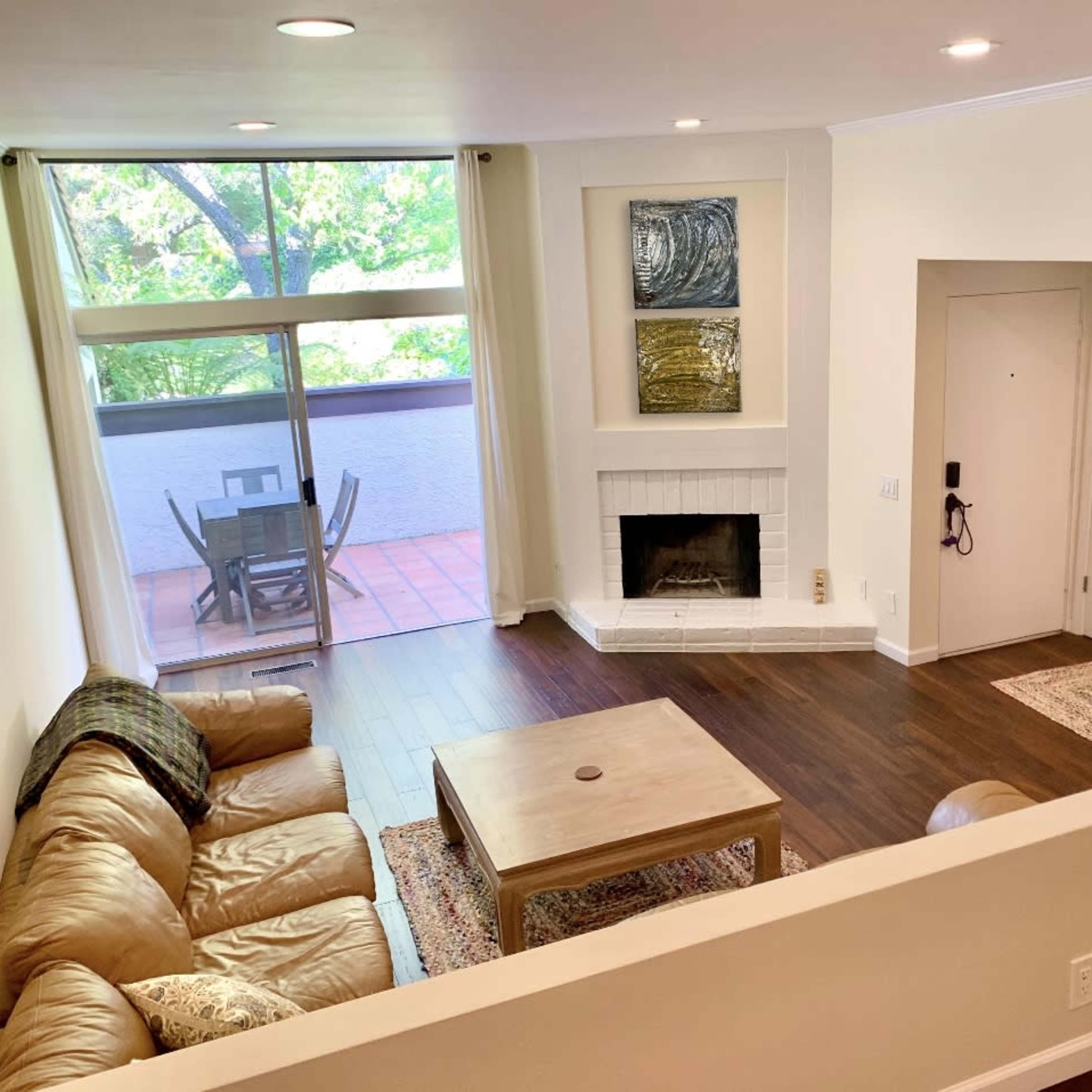 A living room with a tan leather sofa, a wooden coffee table, and a fireplace, adjacent to a dining area with a patio view.